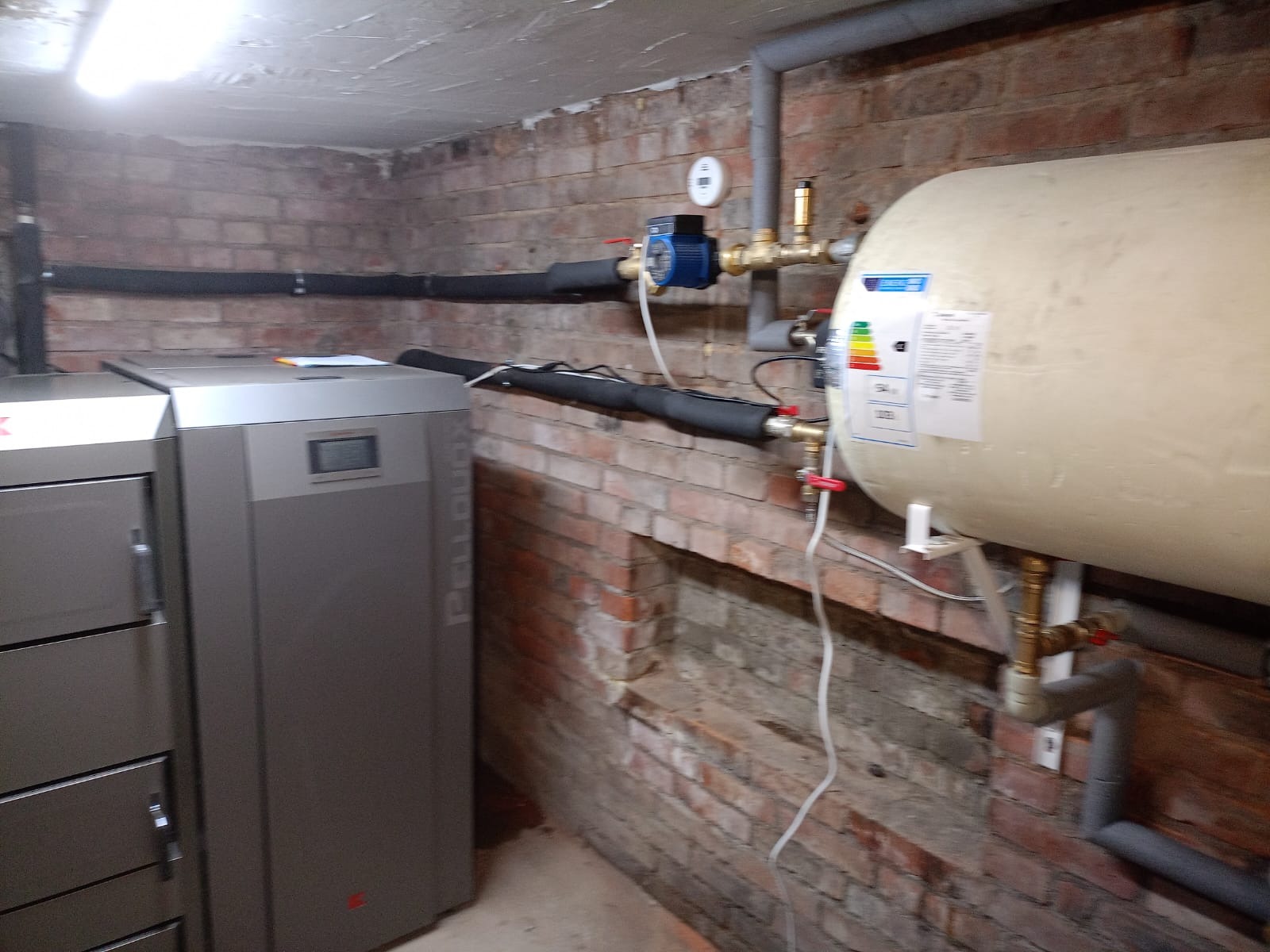 Szary piec na paliwo stałe Pelloux, zbiornik akumulacyjny i instalacja CO na tle ceglanej ściany w piwnicy. Widoczne rury i zawory.