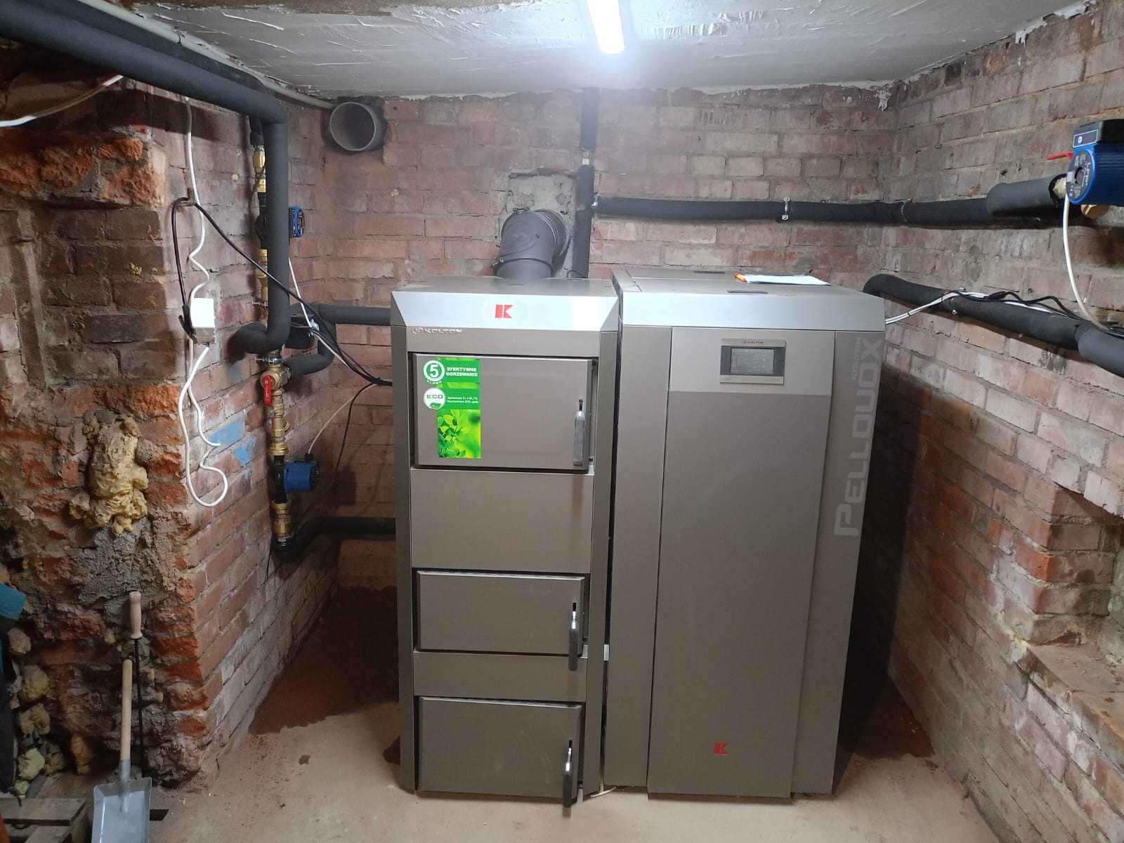 Nowoczesny piec na pellet Kolton PellDuoX w kotłowni z instalacją CO, widoczne rury i ceglana ściana. Piec z naklejką '5 lat efektywne ogrzewanie'.