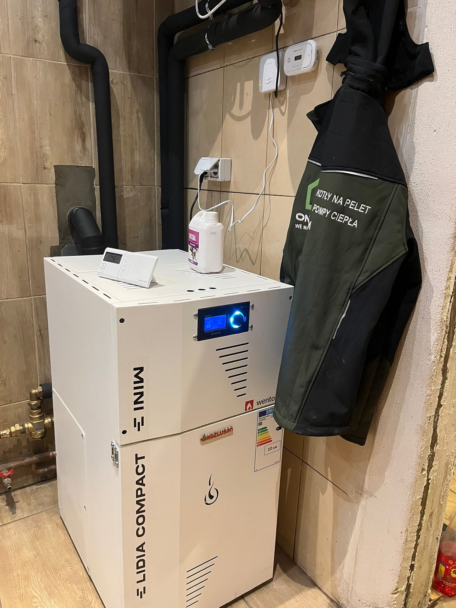 Biały piec na pellet Lidia Compact Mini z czarnymi rurami i kurtką z napisem 'Kotły na Pelet Pompy Ciepła' w tle, zamontowany w pomieszczeniu z beżowymi płytkami.