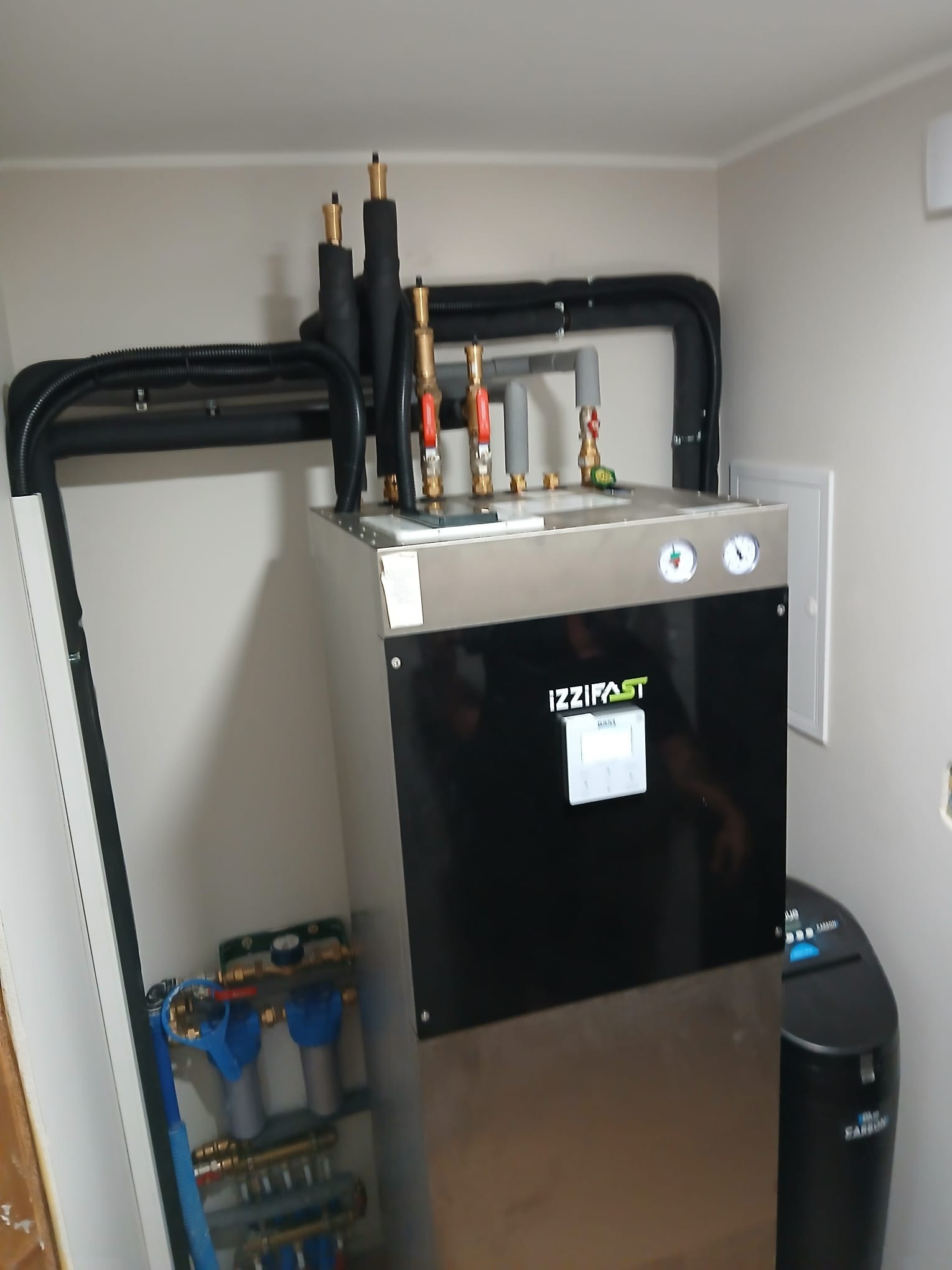 Nowoczesny piec Izzifast z kompletną instalacją hydrauliczną i rurami w izolacji, zamontowany w pomieszczeniu. Widoczne filtry i zawory.