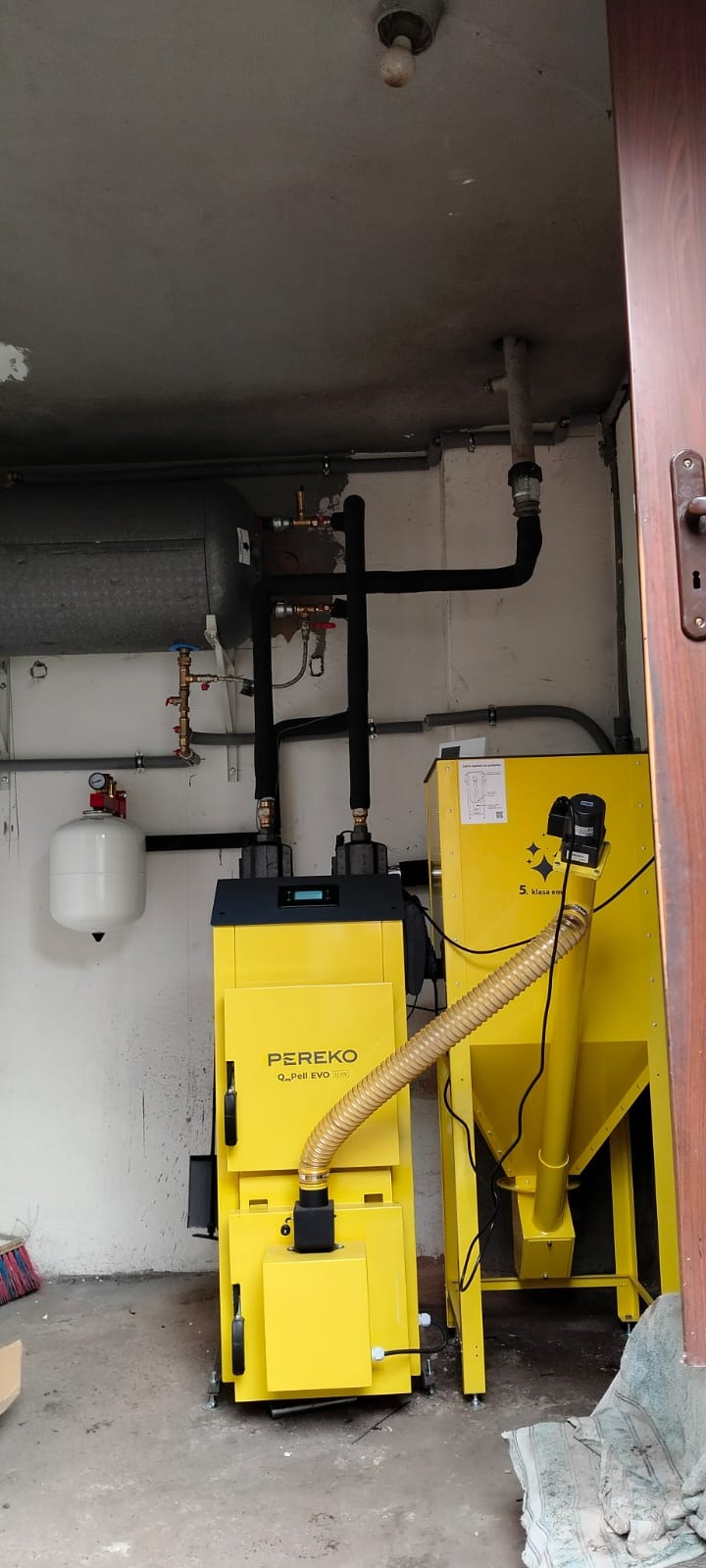 Żółty piec na pellet marki PEREKO z podajnikiem, zainstalowany w kotłowni. Widoczne rury instalacji CO, zbiornik wyrównawczy i oświetlenie sufitowe.