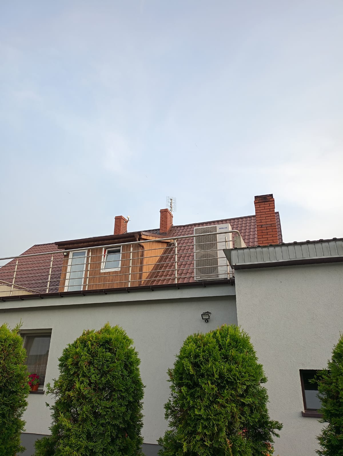 Biała pompa ciepła Samsung na dachu domu z brązową dachówką, widoczna z poziomu ogrodu z trzema tujami. Czerwona cegła komina i antena TV.