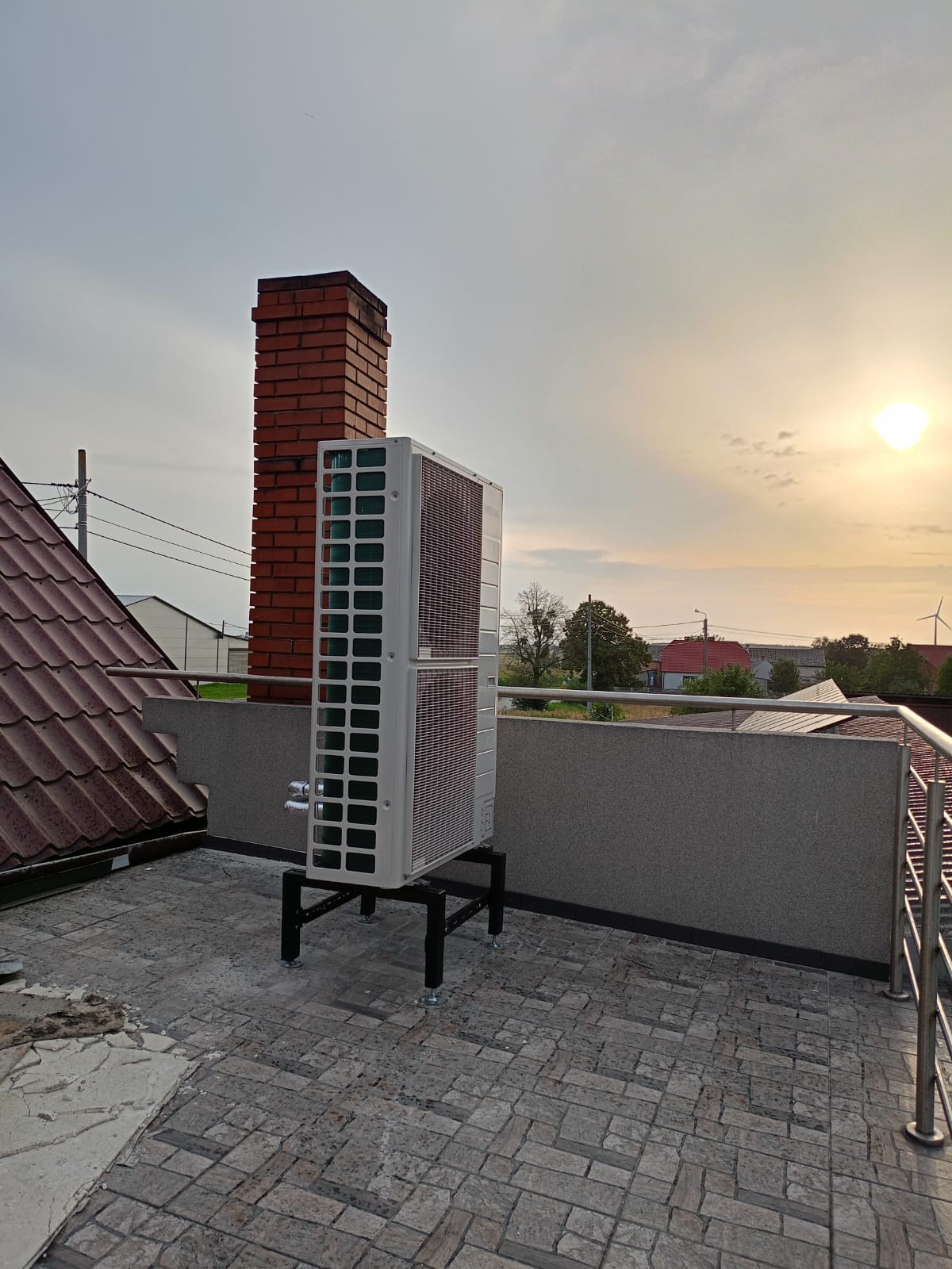 Biała pompa ciepła na dachu budynku, z ceglanym kominem w tle i zachodzącym słońcem. Widoczna instalacja na czarnym stelażu, balustrada i fragment dachu z dachówką.