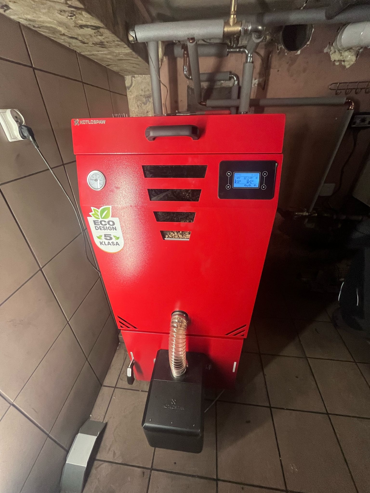 Czerwony piec na paliwo stałe marki Kotłospaw z widocznym panelem sterowania, naklejką Eco Design i elementami instalacji w tle, umieszczony w pomieszczeniu z brązowymi płytkami.