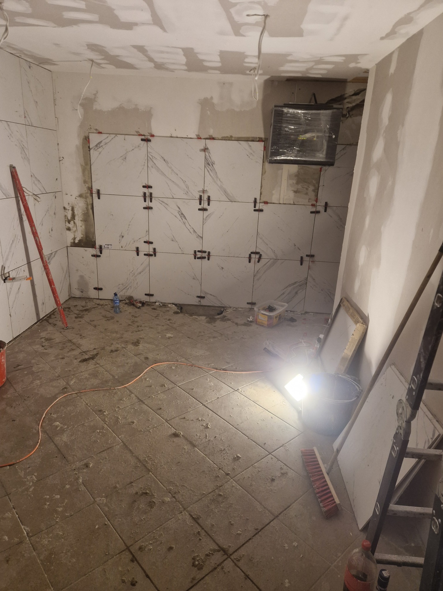Wnętrze w trakcie remontu: białe płytki imitujące marmur na ścianach, krzyżyki dystansowe, surowy sufit, narzędzia i drabina. Widoczne ślady prac remontowych na podłodze.