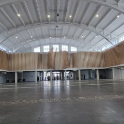 ŁYSZYK GROUP SPÓŁKA Z OGRANICZONĄ ODPOWIEDZIALNOŚCIĄ - Przestronne wnętrze hali z drewnianą dekoracją ścienną z pionowych listew o falistym kształcie, widoczne prace wykończeniowe, malowanie ścian, osoby w tle.