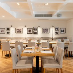 ŁYSZYK GROUP SPÓŁKA Z OGRANICZONĄ ODPOWIEDZIALNOŚCIĄ - Jasne wnętrze restauracji w Poznaniu po remoncie, z parkietem w jodełkę, białymi ścianami ozdobionymi czarno-białymi zdjęciami w ramkach, drewnianymi stolikami z nakryciem i beżowymi krzesłami.