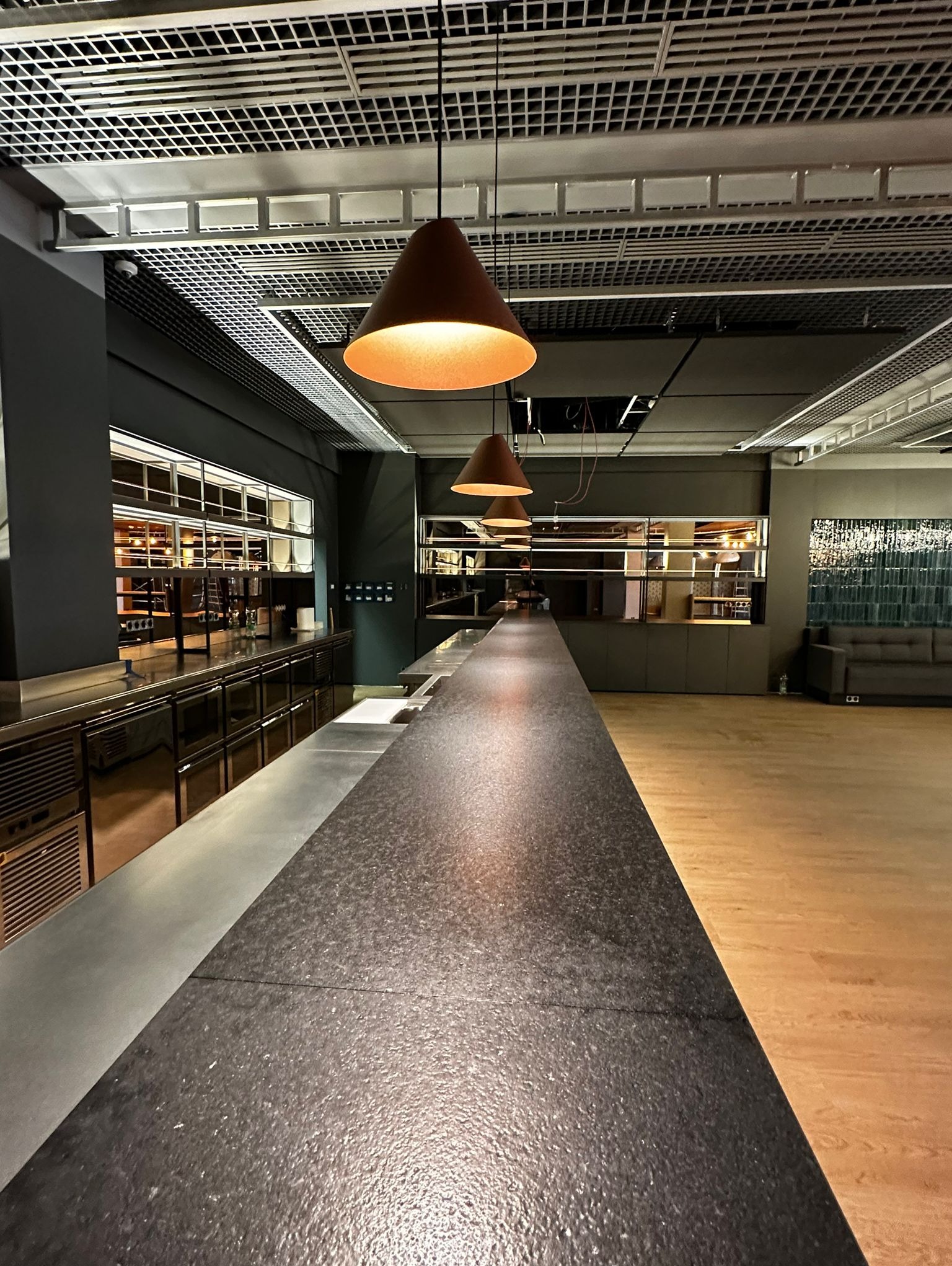 Długa, ciemna lada barowa z metalowymi szafkami pod spodem, oświetlona trzema stożkowymi lampami wiszącymi, widok na przestronne, puste wnętrze z drewnianą podłogą i kratownicowym sufitem.