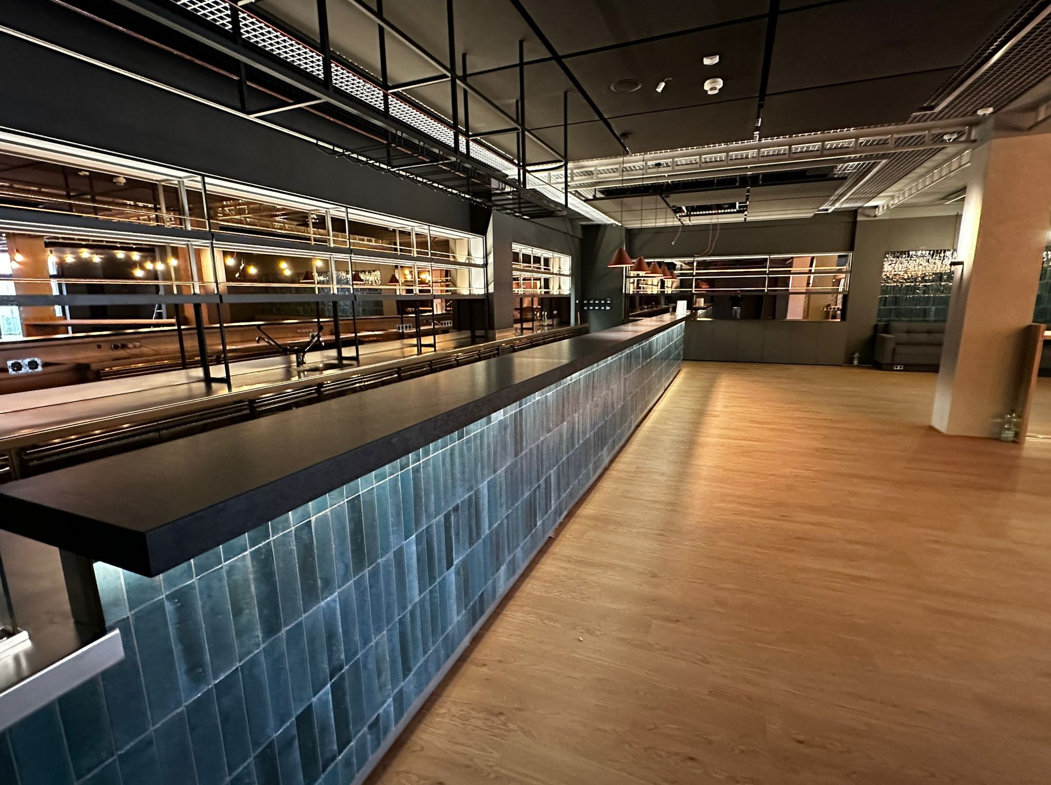 Długi bar wykończony pionowymi, turkusowymi płytkami ceramicznymi w nowoczesnym wnętrzu biurowym z drewnianą podłogą i metalową konstrukcją sufitową.