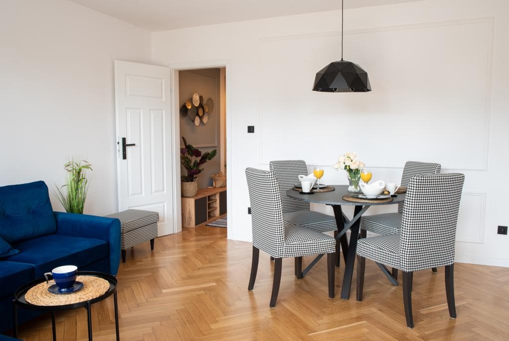 Jasne wnętrze mieszkania z parkietem w jodełkę, okrągłym stołem z krzesłami w pepitkę, niebieską sofą i dekoracjami na ścianie w tle.