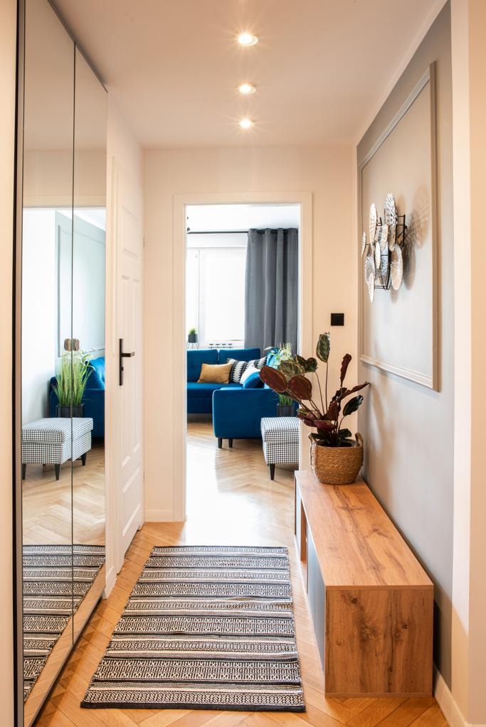 Nowoczesny przedpokój z lustrzaną szafą, drewnianą konsolą z rośliną w koszyku i dywanem w geometryczne wzory, widok na salon z niebieską sofą.