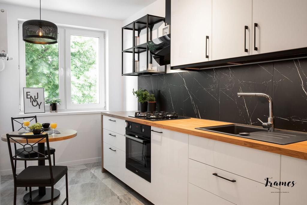 Jasna kuchnia z białymi szafkami, drewnianym blatem i czarnym marmurowym backsplashiem, okrągły stół z czarnymi krzesłami i napojami przy oknie.