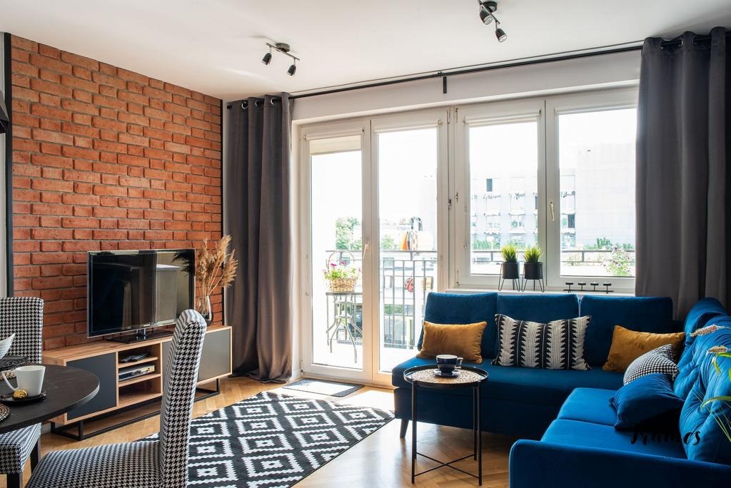 Salon z ceglaną ścianą, niebieską sofą narożną, czarno-białym dywanem i wyjściem na balkon z widokiem na zieleń.