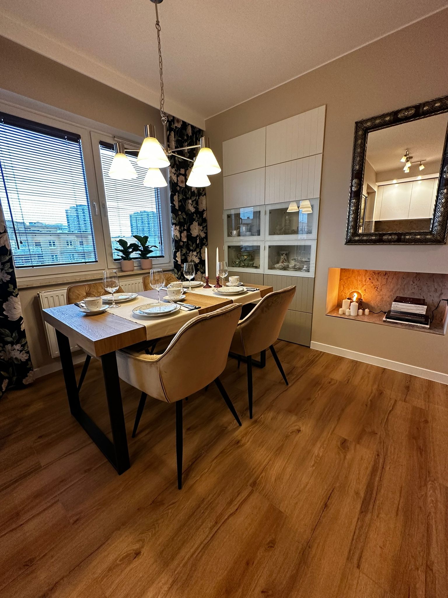 Ustawiony stół w jadalni z drewnianym blatem i czarnymi nogami, beżowe krzesła, okno z żaluzjami, dekoracyjne zasłony w kwiaty, zabudowa meblowa z przeszklonymi witrynami, lustro w ozdobnej ramie...