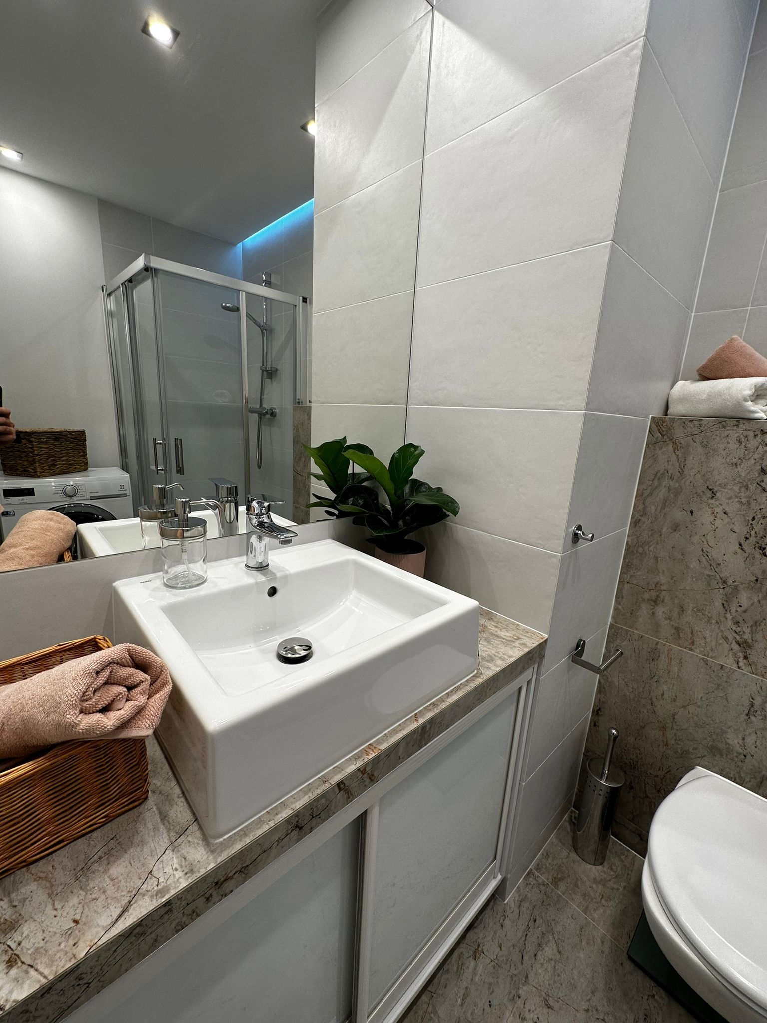 Nowoczesna łazienka z białą, prostokątną umywalką nablatową, baterią chromowaną, rośliną doniczkową, szarymi płytkami ściennymi, marmurowym blatem i kabiną prysznicową w tle.