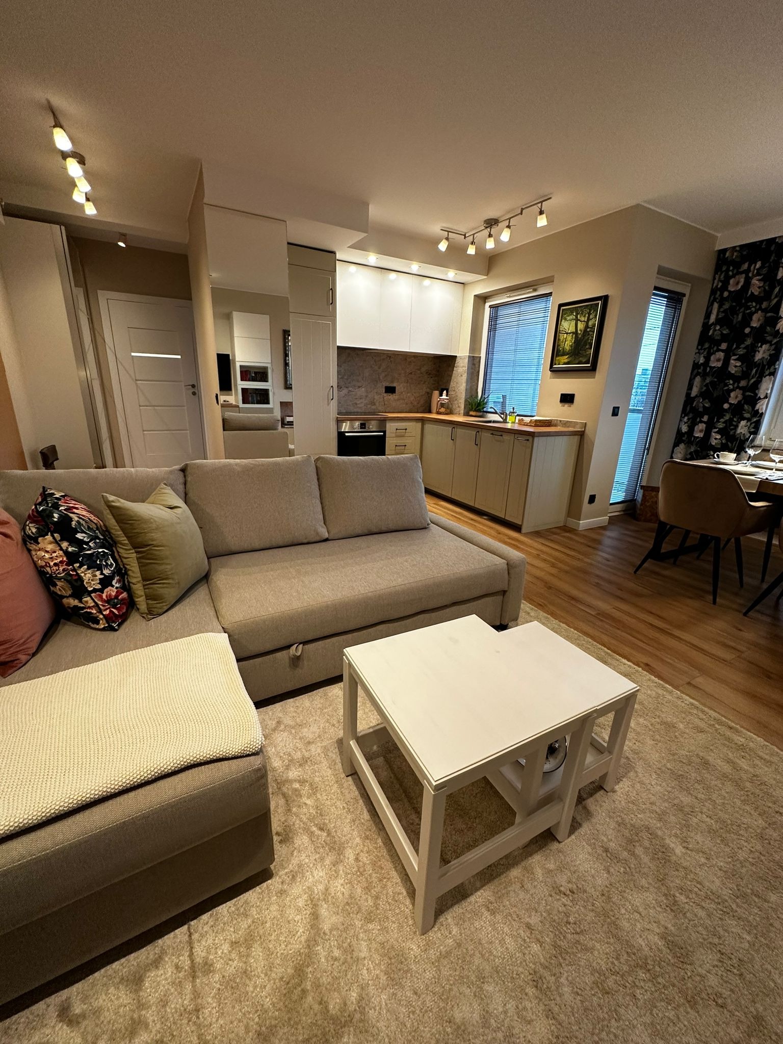 Jasne wnętrze mieszkania z aneksem kuchennym, widoczny narożnik z poduszkami w kwiaty, biały stolik kawowy i drewniana podłoga.