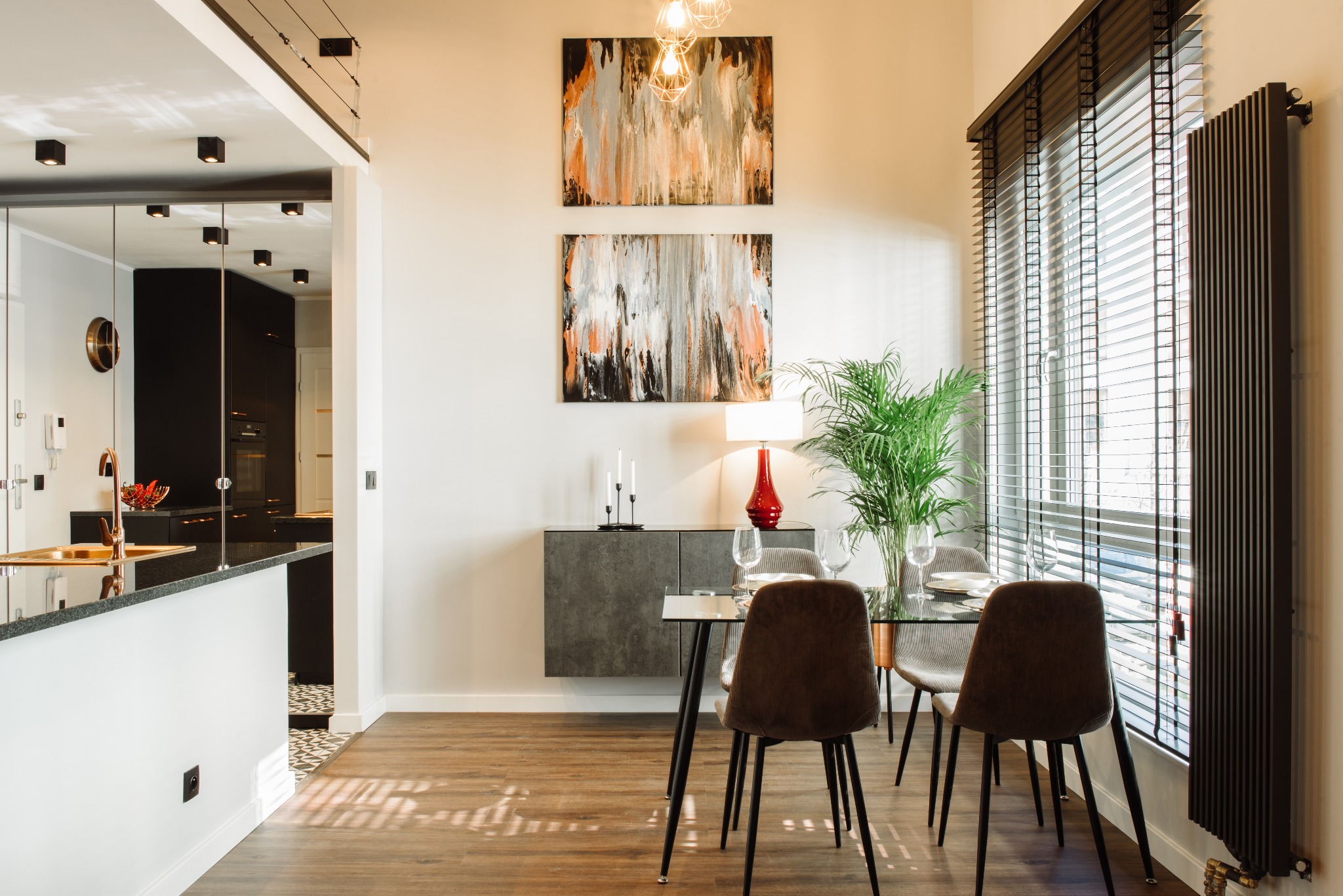 Jasne, nowoczesne wnętrze: jadalnia z szklanym stołem, krzesłami obitymi brązowym sztruksem, szarą szafką podwieszaną, abstrakcyjnymi obrazami na ścianie i wysokim grzejnikiem w kolorze grafitowym...