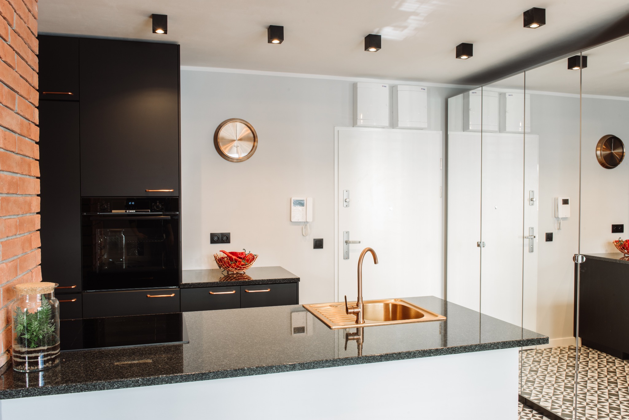 Nowoczesna kuchnia z czarnymi szafkami, miedzianymi uchwytami i armaturą, granitowym blatem i ceglaną ścianą w tle, oświetlona punktowymi lampami sufitowymi.