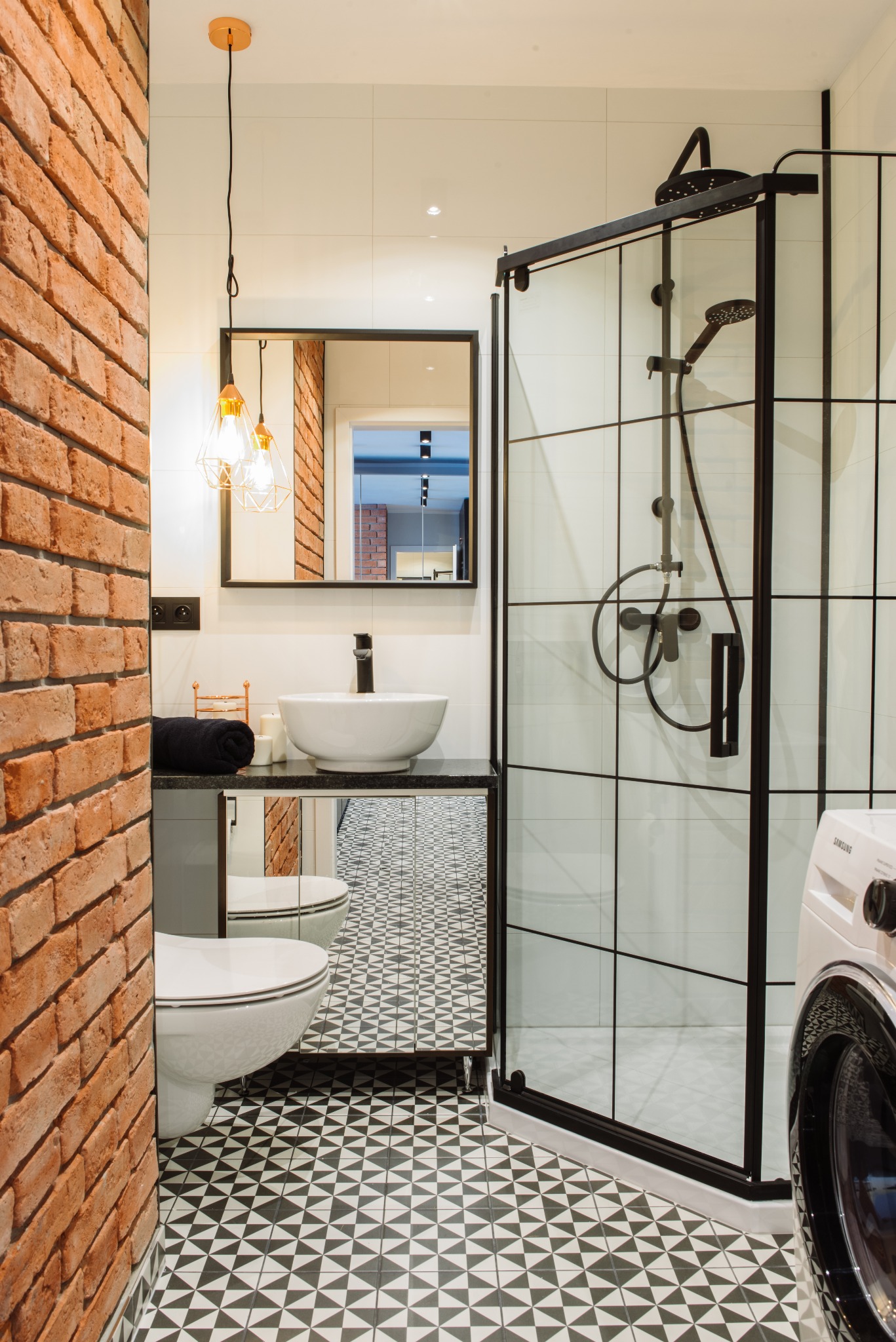 Nowoczesna łazienka z ceglaną ścianą, kabiną prysznicową typu walk-in z czarnymi profilami, umywalką nablatową na szafce z lustrzanymi frontami i wzorzystą podłogą.