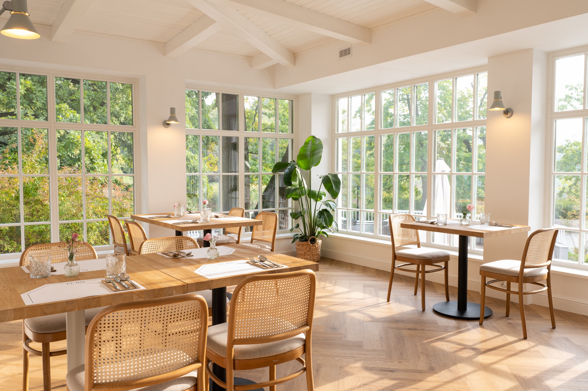 Jasne wnętrze restauracji z drewnianymi stołami, rattanowymi krzesłami i dużymi oknami z widokiem na zieleń, parkiet ułożony w jodełkę, na stołach nakrycia i wazoniki z różowymi kwiatami.