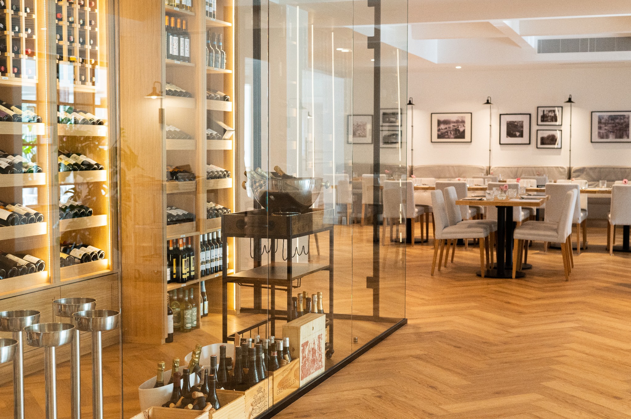 Eleganckie wnętrze restauracji z drewnianą podłogą w jodełkę, szklaną ścianą, regałami z winami i nakrytymi stolikami.