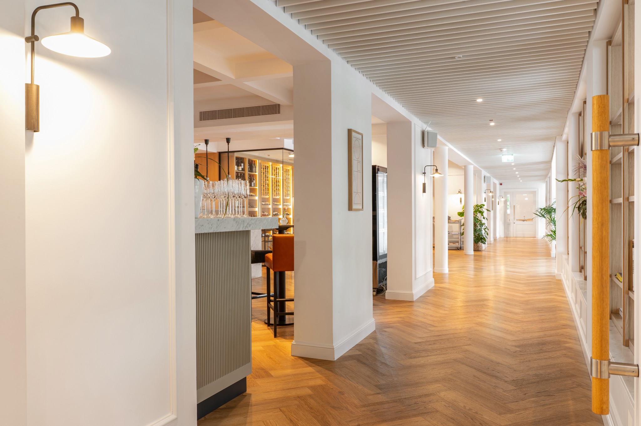 Długi korytarz w hotelu z parkietem ułożonym w jodełkę, białymi ścianami i kolumnami, widok na bar z wysokimi szklankami i butelkami w tle, oświetlony lampami ściennymi i punktowymi na suficie.