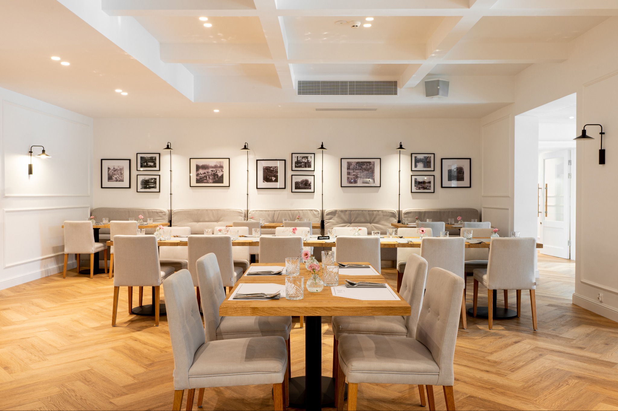 Jasne wnętrze restauracji w Poznaniu po remoncie, z parkietem w jodełkę, białymi ścianami ozdobionymi czarno-białymi zdjęciami w ramkach, drewnianymi stolikami z nakryciem i beżowymi krzesłami.