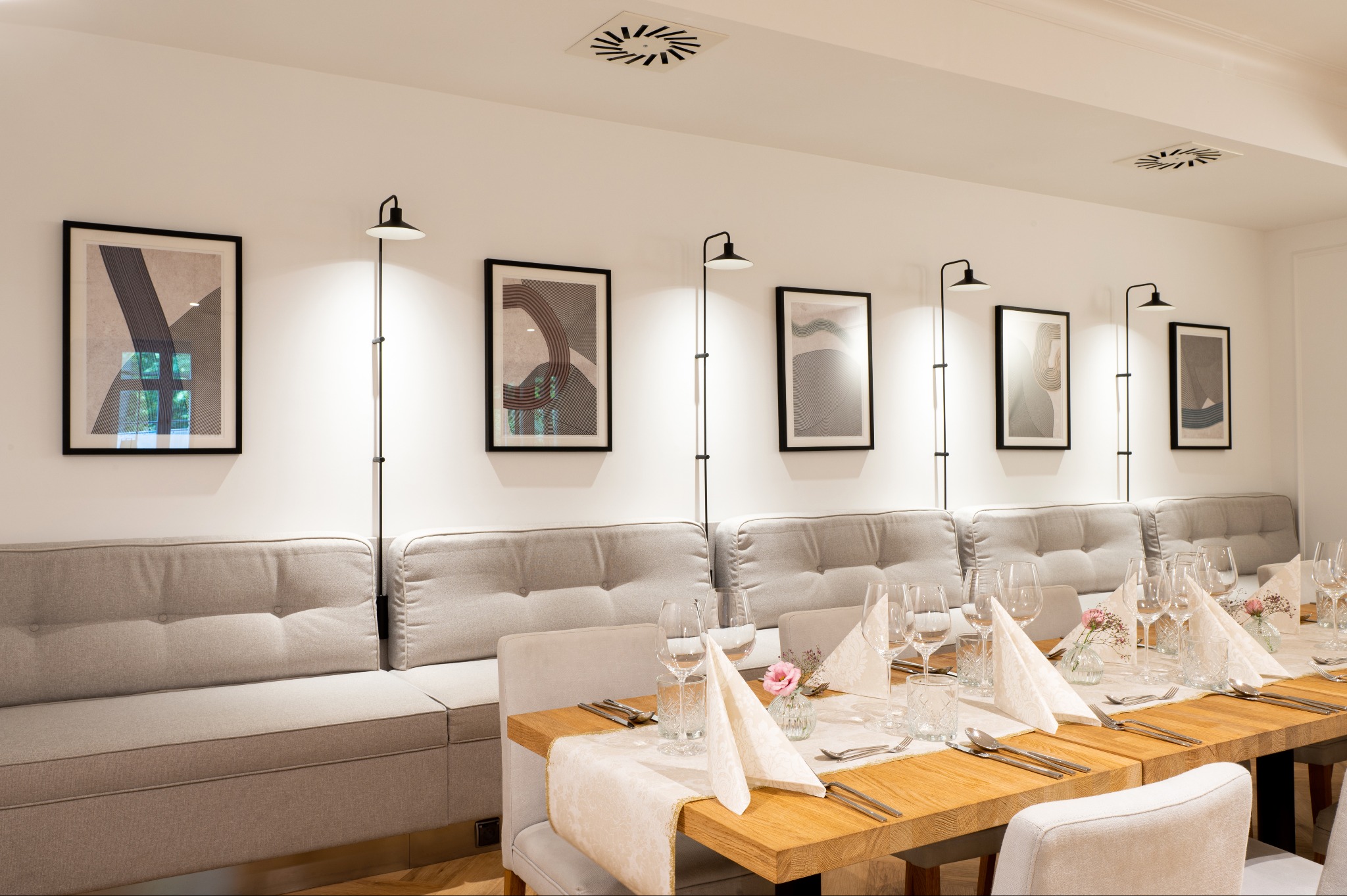 Jasne wnętrze restauracji z długim drewnianym stołem nakrytym dla gości, szarym narożnikiem z tapicerowanymi siedziskami i oparciami, oraz minimalistycznymi obrazami na ścianie oświetlonymi przez...