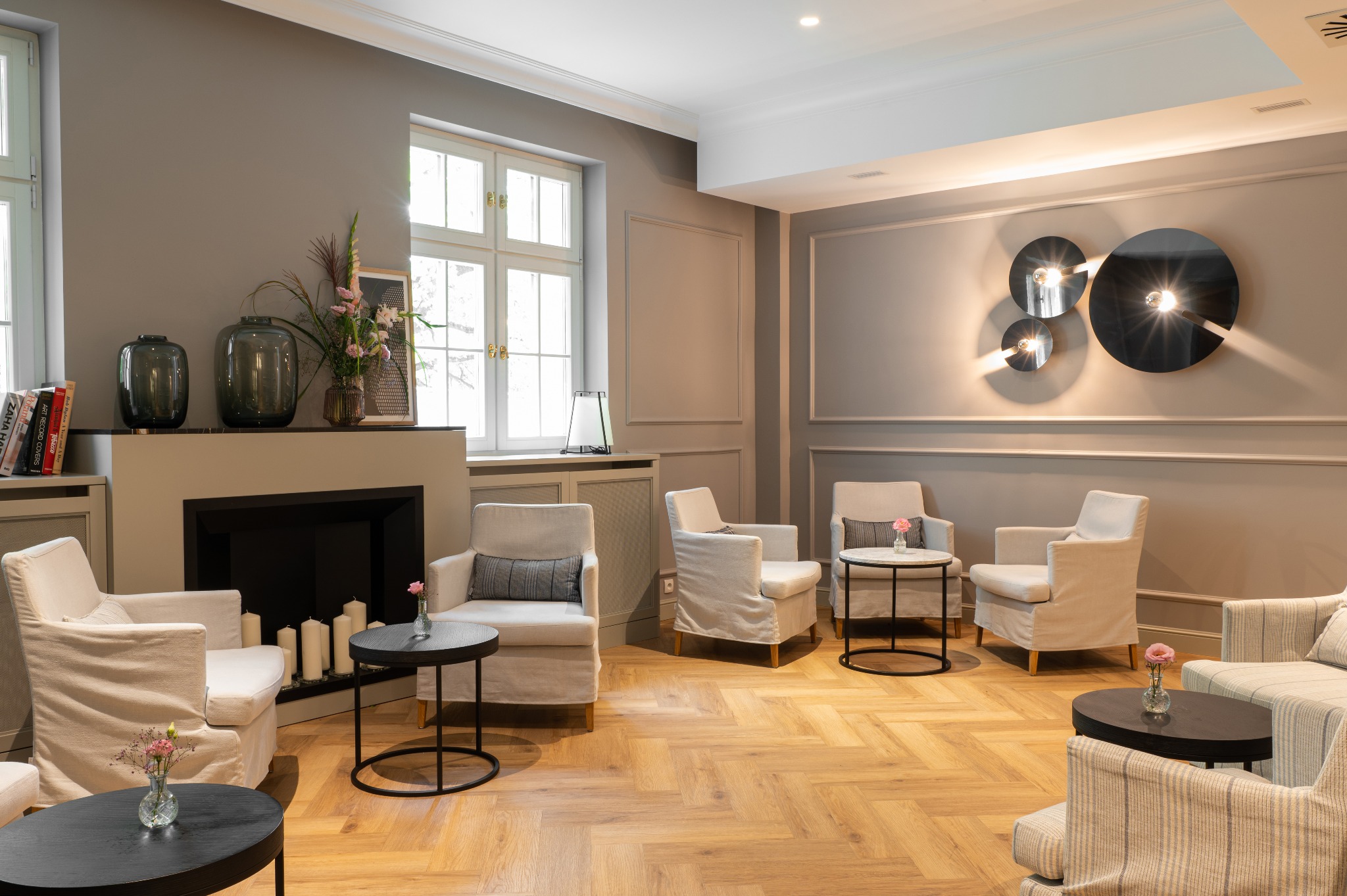 Eleganckie wnętrze salonu z parkietem w jodełkę, kremowymi fotelami i dekoracyjnym kominkiem z świecami, oświetlone nowoczesnymi lampami.