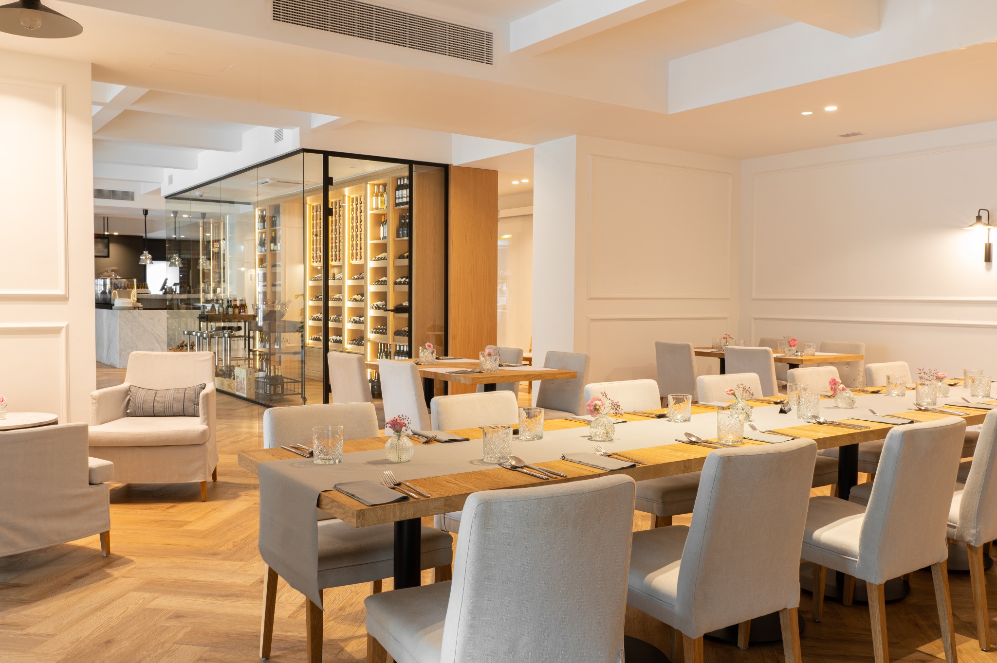 Jasne, przestronne wnętrze restauracji z parkietem w jodełkę, drewnianymi stołami nakrytymi szarymi bieżnikami, szarymi krzesłami i szklanymi wazonikami z różowymi kwiatami. W tle przeszklona...