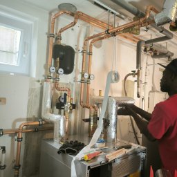 Elektro-S - Instalator w czerwonej koszulce owija izolacją termiczną miedziane rury grzewcze w kotłowni, widoczne narzędzia i mierniki na kotle grzewczym.