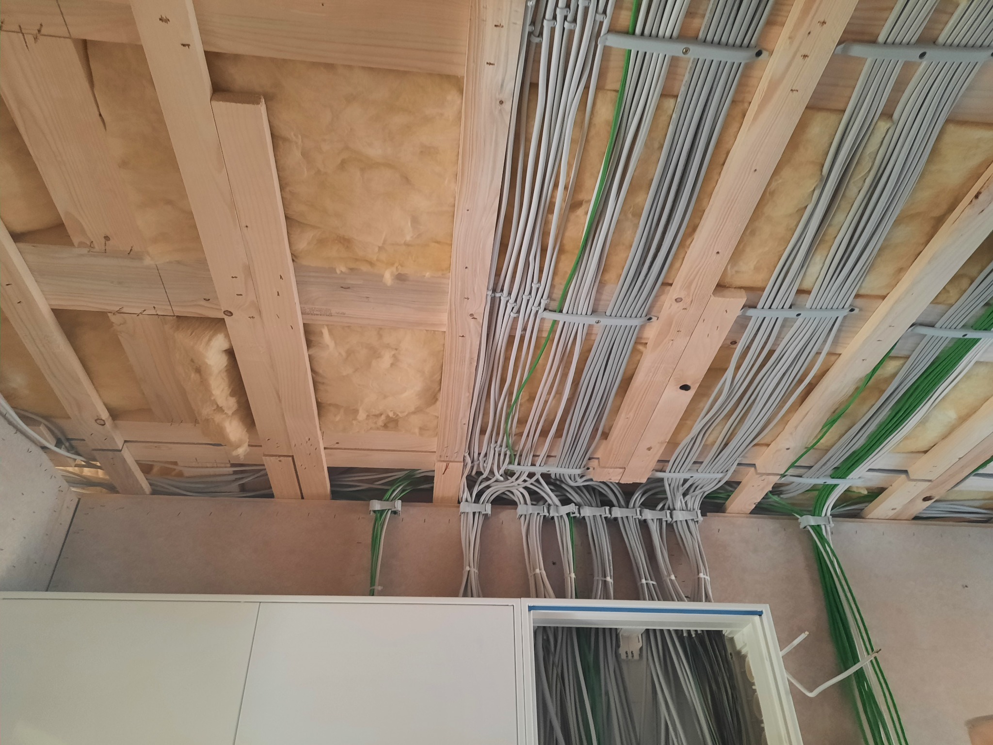 Szczegółowe ujęcie instalacji elektrycznej pod stropem, widoczne liczne szare przewody prowadzone w korytkach, przeplatane zielonymi kablami, strop ocieplony wełną mineralną, fragment szafki...