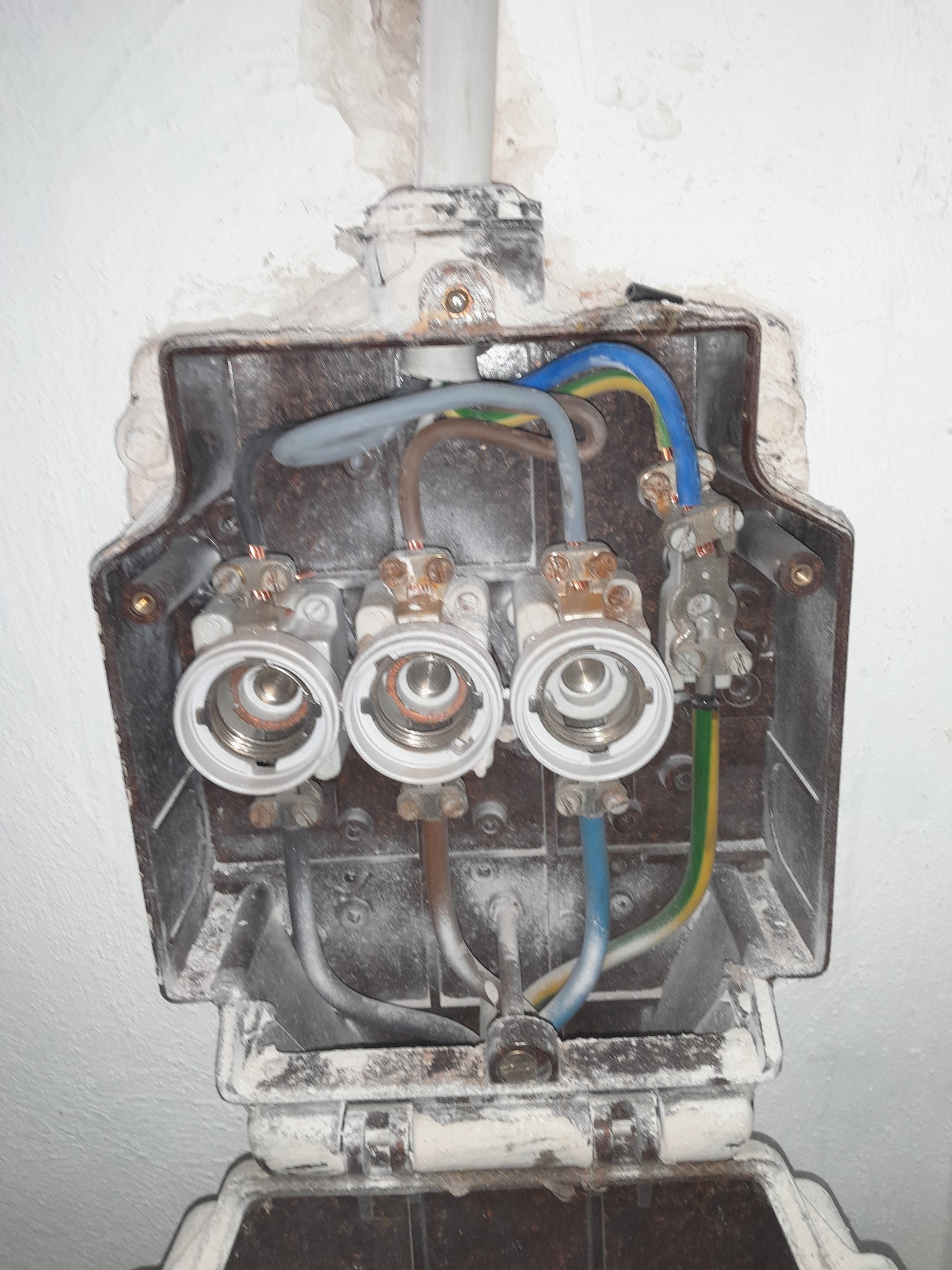 Otwarta, stara skrzynka bezpiecznikowa z trzema gniazdami na bezpieczniki i widocznymi kablami w kolorach niebieskim, żółto-zielonym i szarym, zamontowana na białej ścianie z ubytkami tynku.