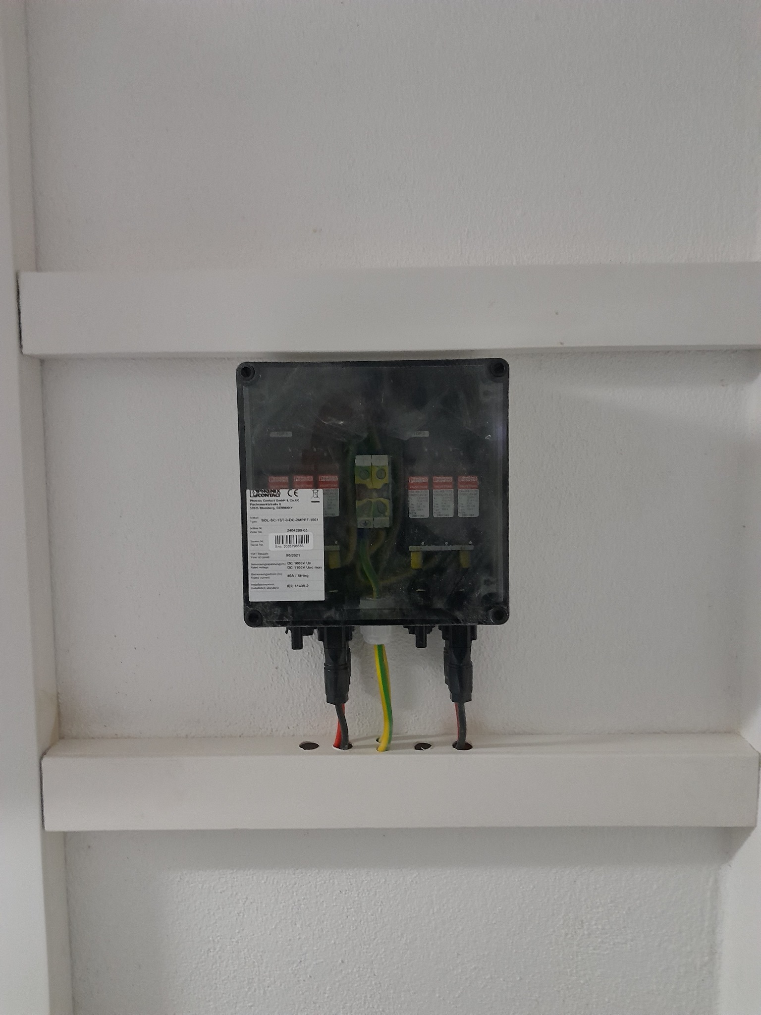 Czarna skrzynka z bezpiecznikami i przewodami elektrycznymi zamontowana na białej ścianie z listwami maskującymi, widoczne naklejki ostrzegawcze i tabliczka znamionowa.