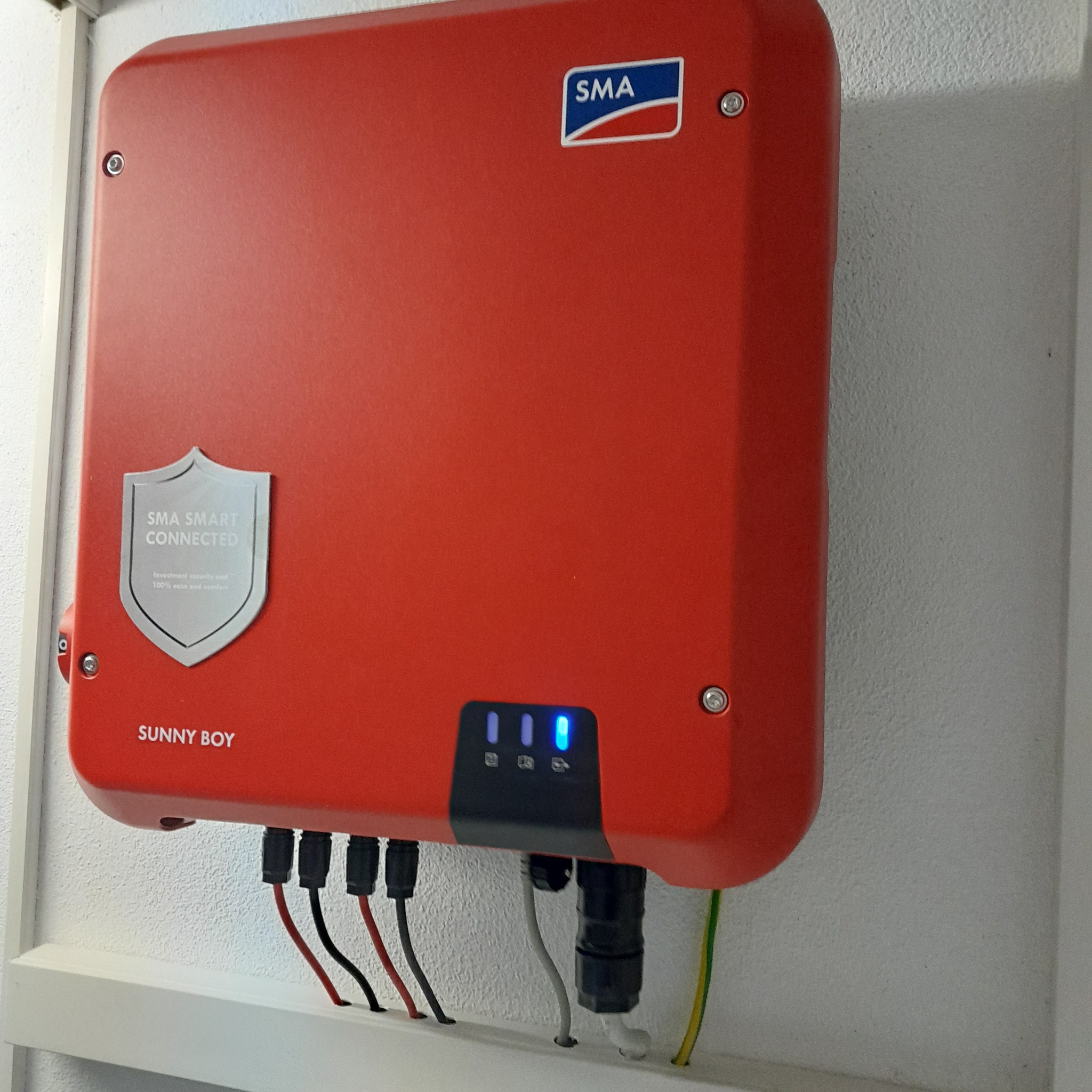 Czerwony inwerter SMA Sunny Boy zamontowany na białej ścianie, widoczne kable i diody sygnalizacyjne.