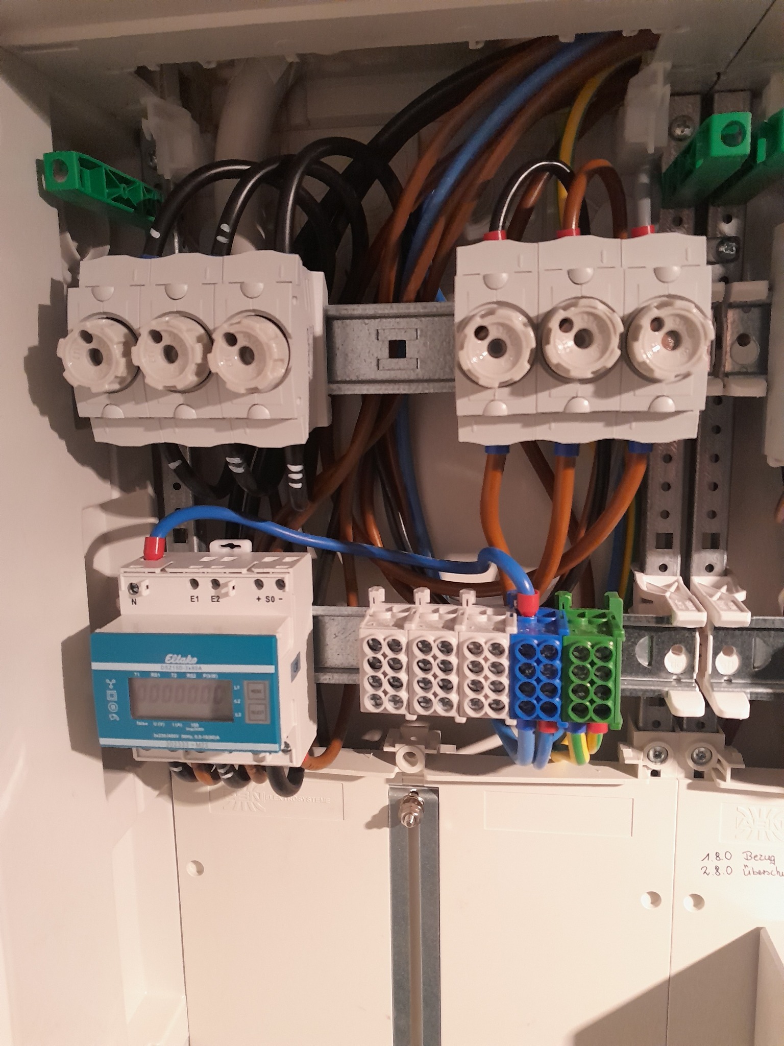 Wnętrze skrzynki elektrycznej z bezpiecznikami topikowymi, licznikiem energii Ellake i kolorowymi przewodami podłączonymi do listew zaciskowych, widoczne napisy na obudowie.