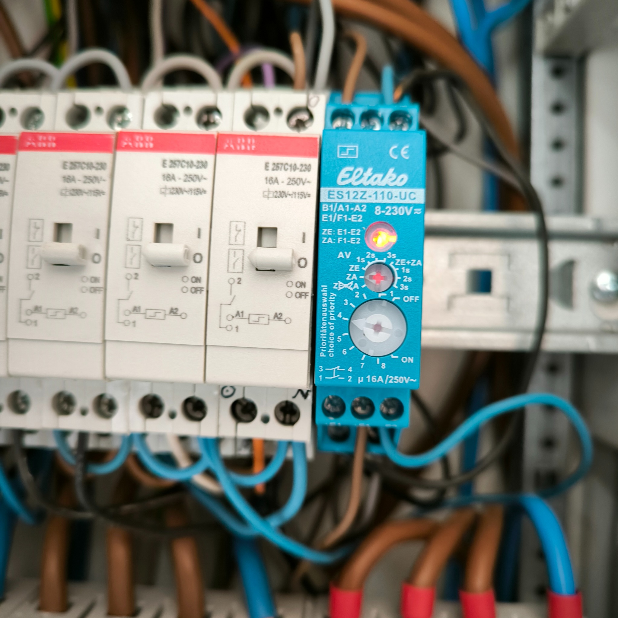 Szczegółowe ujęcie na zabezpieczenia elektryczne w rozdzielnicy, widoczne wyłączniki nadprądowe z oznaczeniami i moduł sterujący Eltako ES12Z-110-UC z ustawionym priorytetem, okablowanie w kolorze...