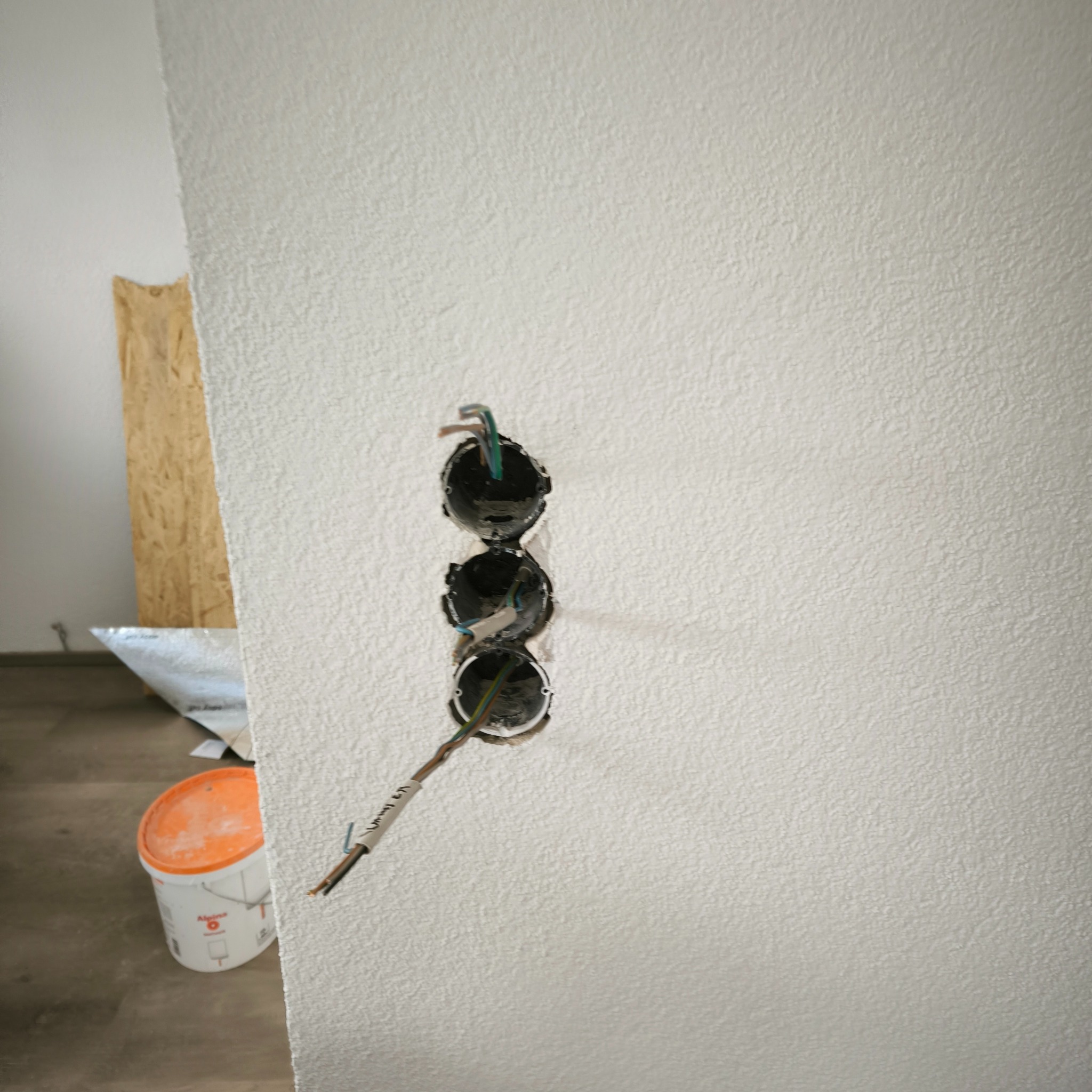 Trzy puste otwory na gniazdka elektryczne w białej, teksturowanej ścianie z wystającymi przewodami, widoczny fragment podłogi i wiadro z farbą w tle.