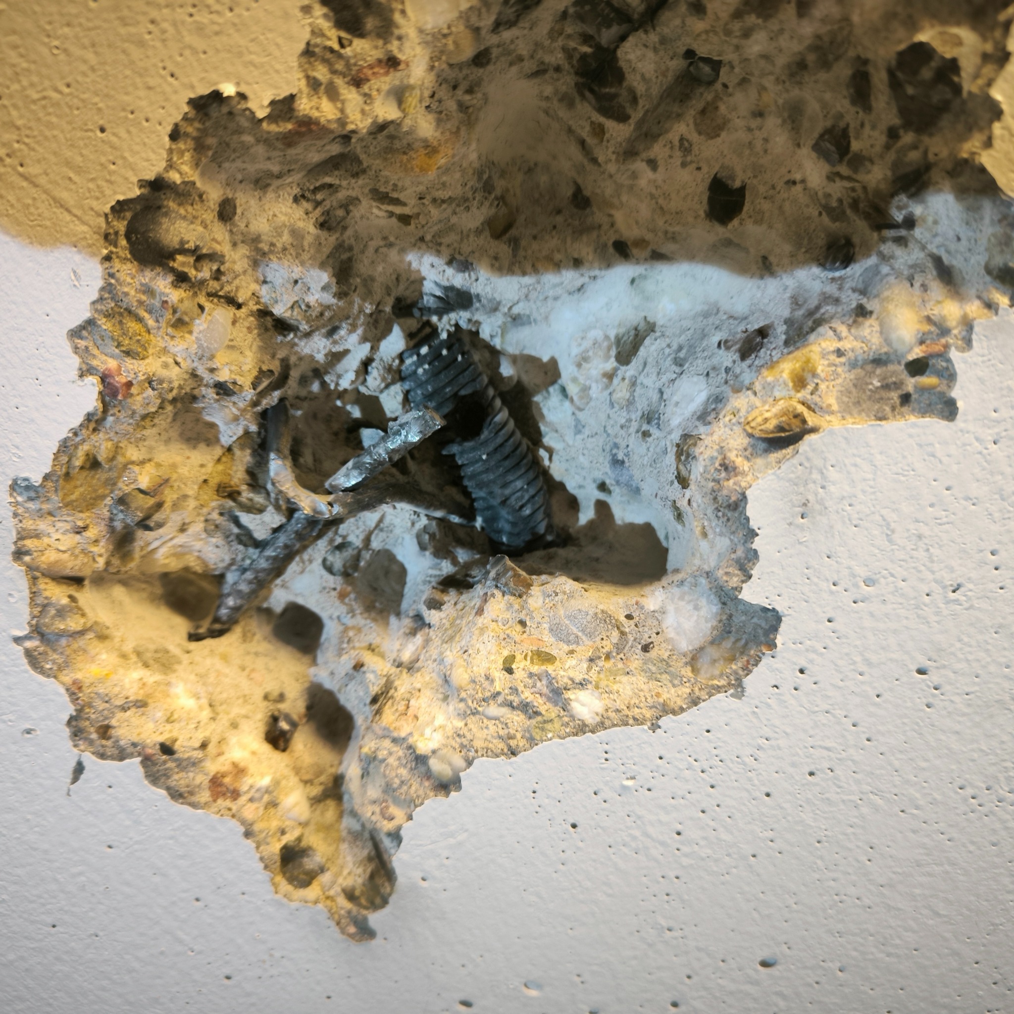 Odsłonięty fragment sufitu z widoczną metalową sprężyną mocującą wewnątrz otworu w betonie, ukazujący warstwy materiału i nierówną powierzchnię.