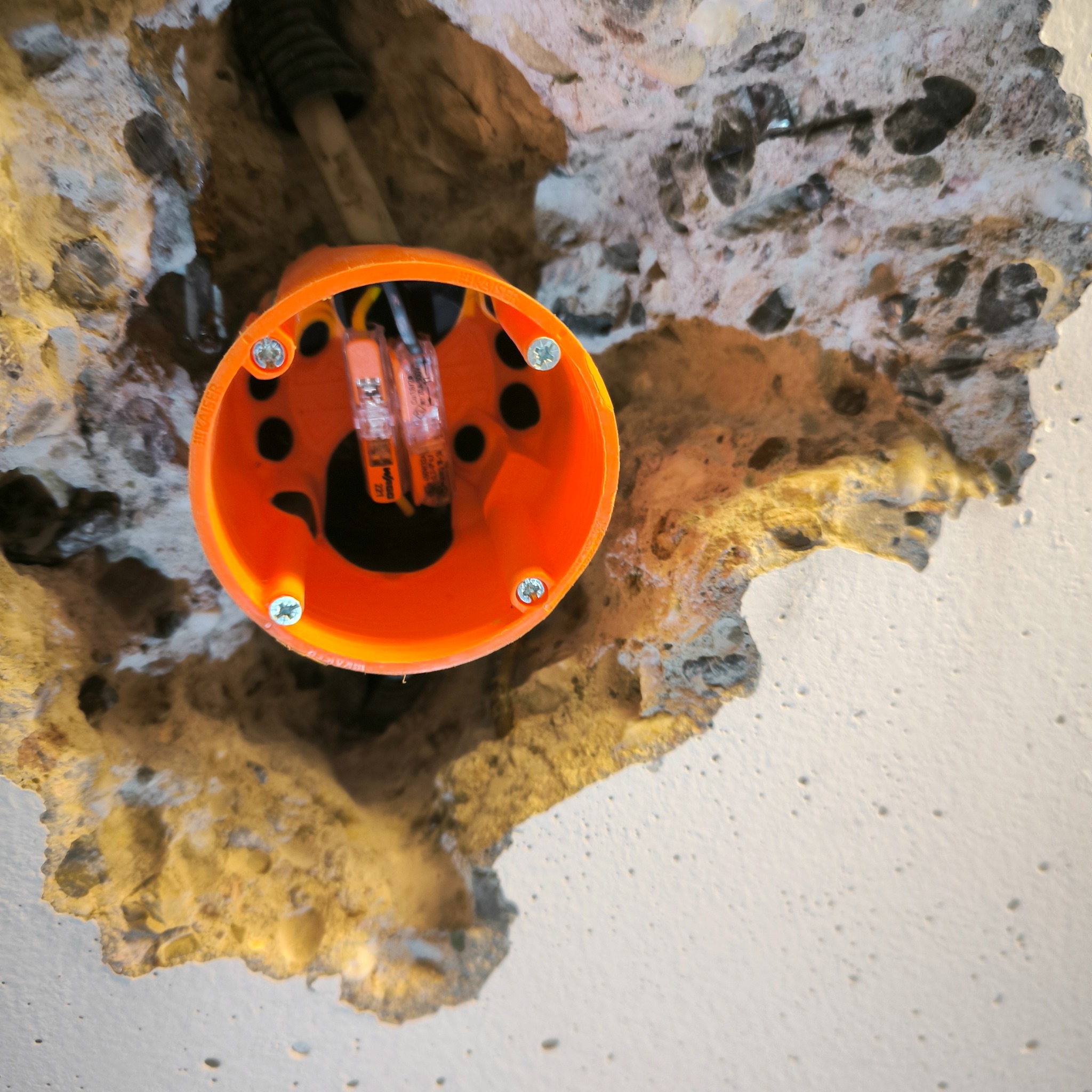 Pomarańczowa puszka elektryczna zamontowana w suficie z widocznymi przewodami i złączkami Wago, otoczona surowym betonem z odpryskami farby.