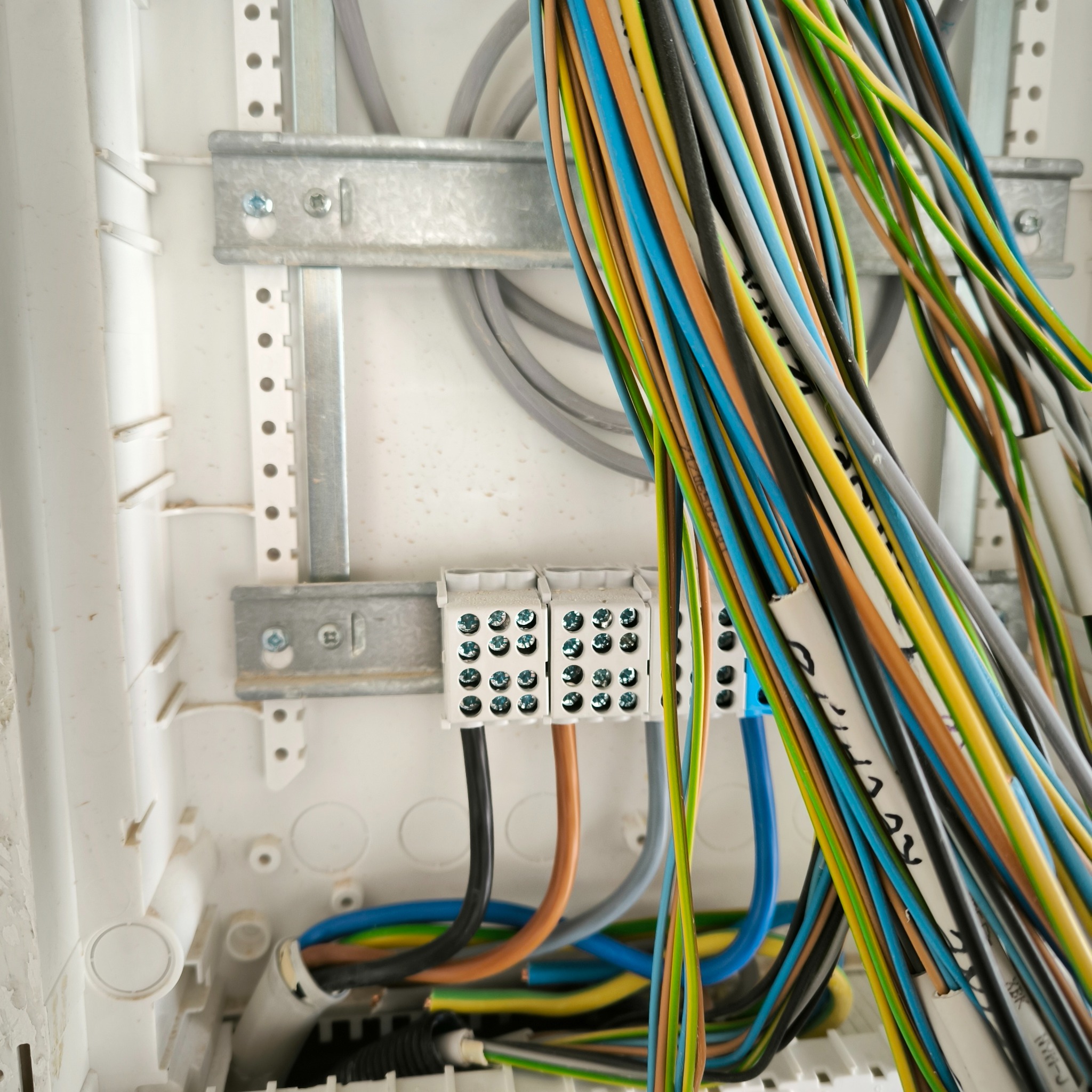 Rozdzielnica elektryczna z podłączonymi przewodami o różnych kolorach, widoczne złącza śrubowe i metalowe szyny montażowe wewnątrz białej obudowy.