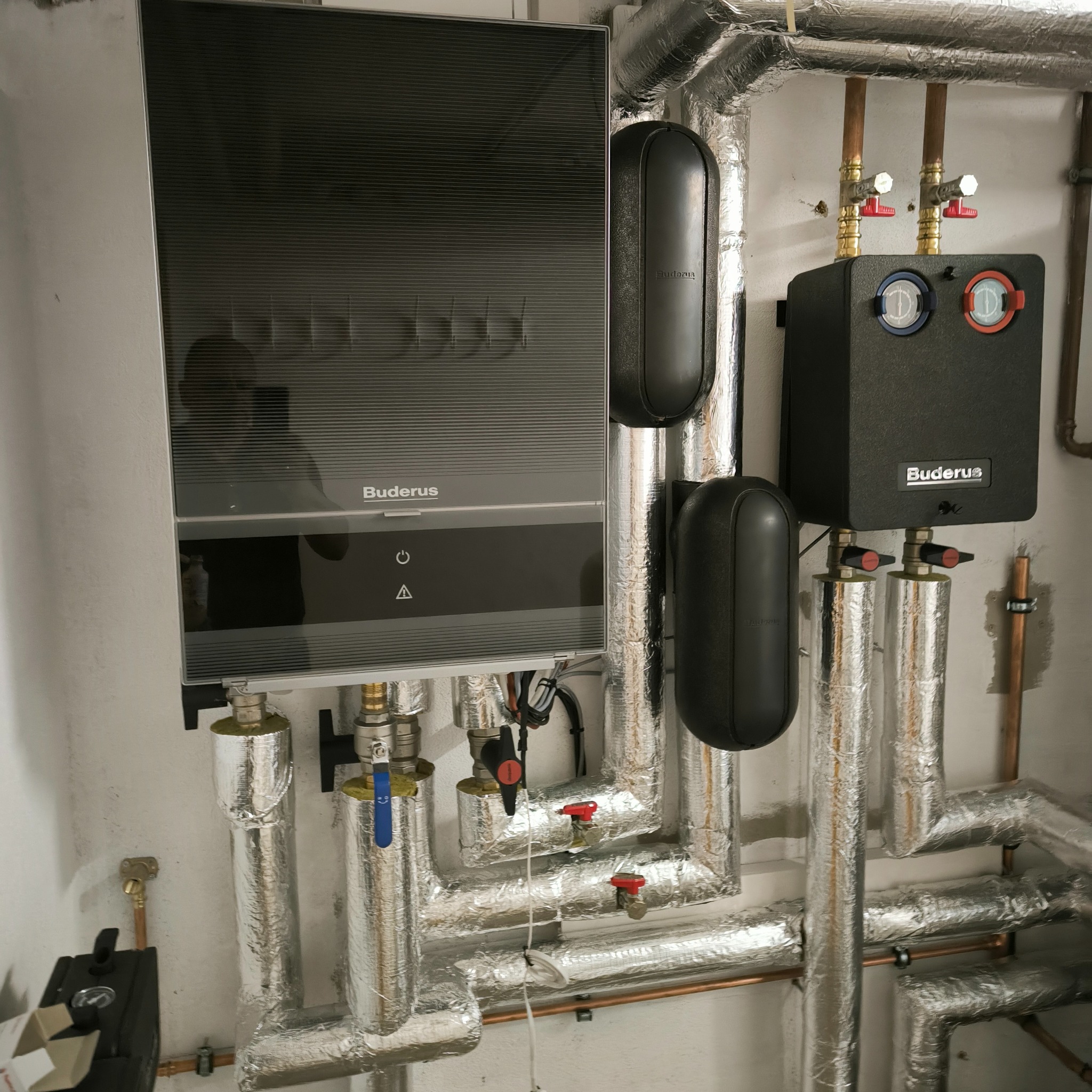 Zainstalowany nowoczesny piec gazowy Buderus z podłączonymi rurami w izolacji aluminiowej, widoczne zawory i elementy sterujące, zamontowane na białej ścianie.