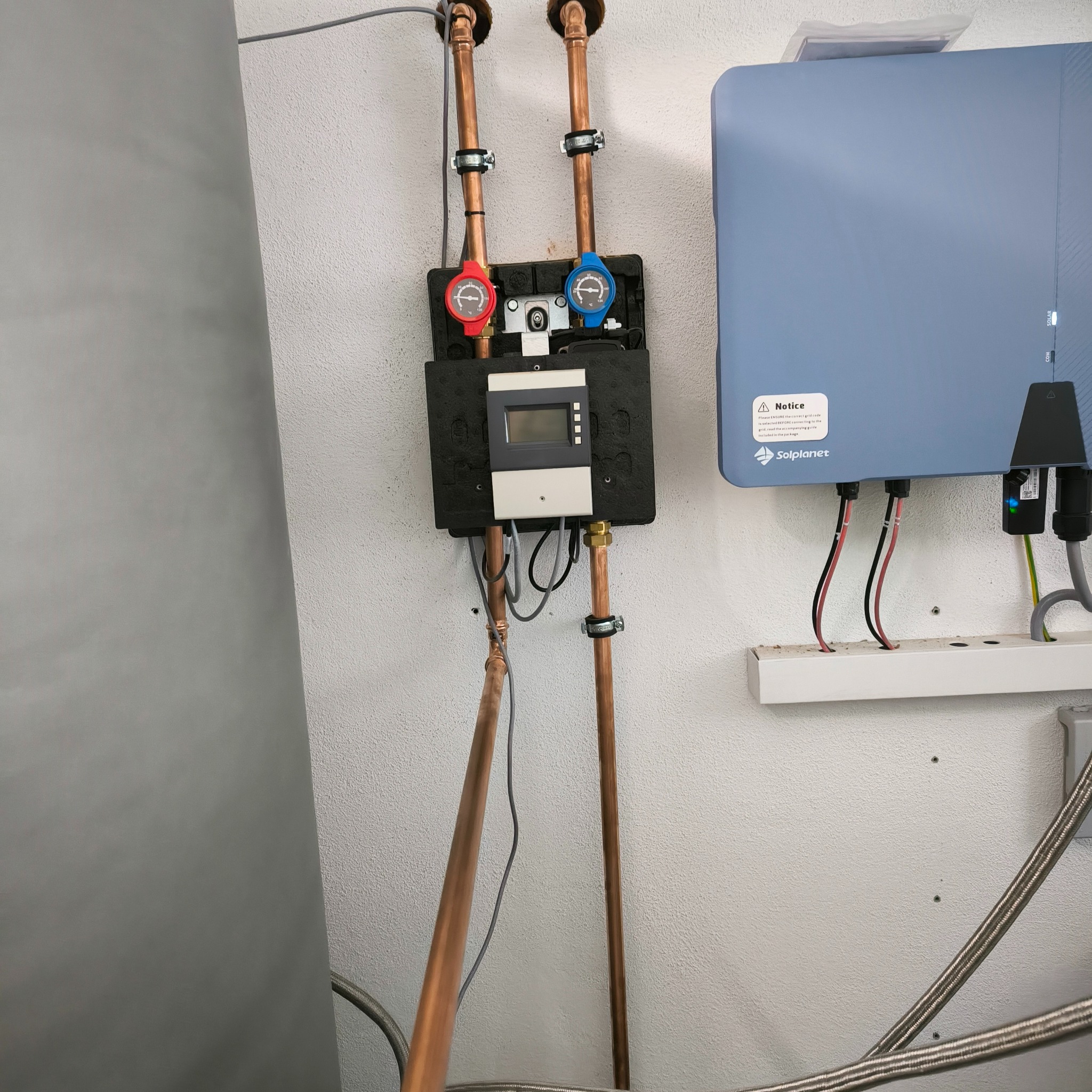 Instalacja solarna z miedzianymi rurami, czujnikami temperatury (czerwony i niebieski) oraz wyświetlaczem, zamontowana na białej ścianie obok szarego zbiornika i niebieskiego falownika Solplanet...
