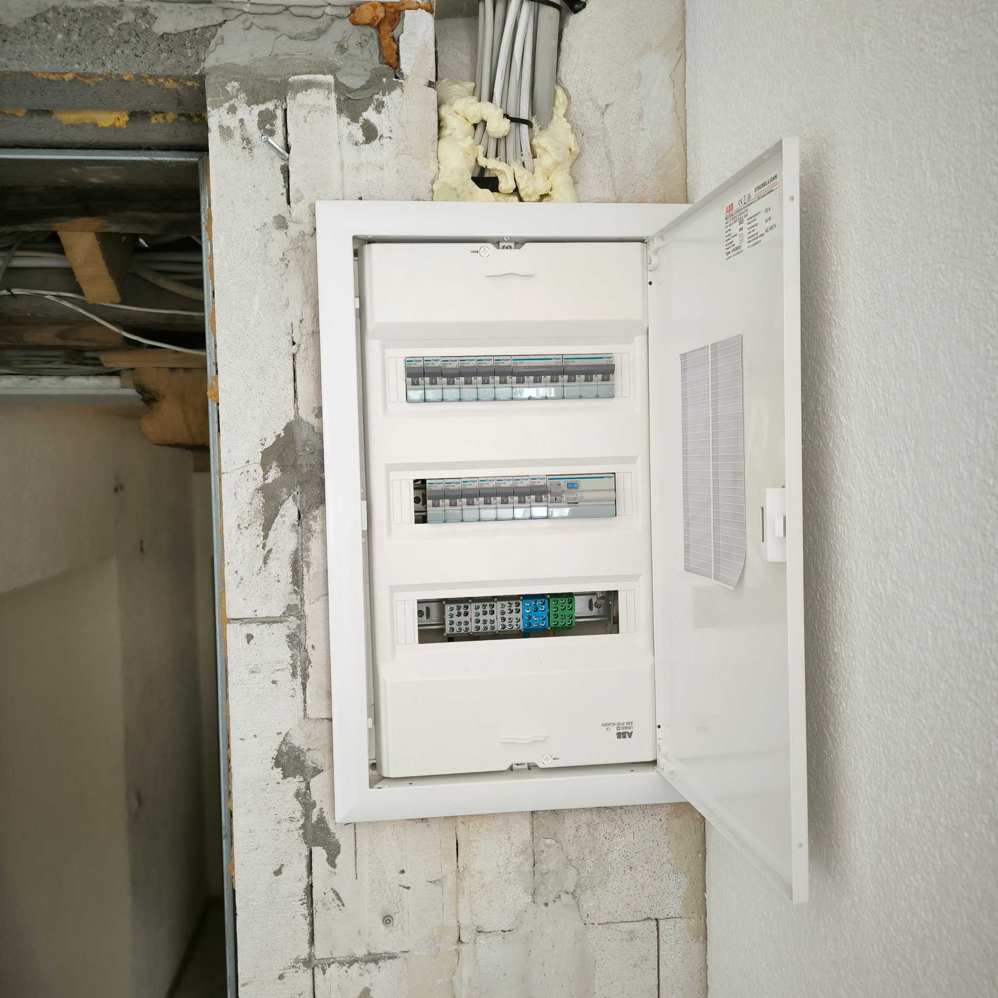 Otwarta skrzynka bezpiecznikowa marki ABB zamontowana w surowej ścianie z betonu komórkowego, widoczne przewody w pianie montażowej u góry i fragment konstrukcji sufitu po lewej stronie.