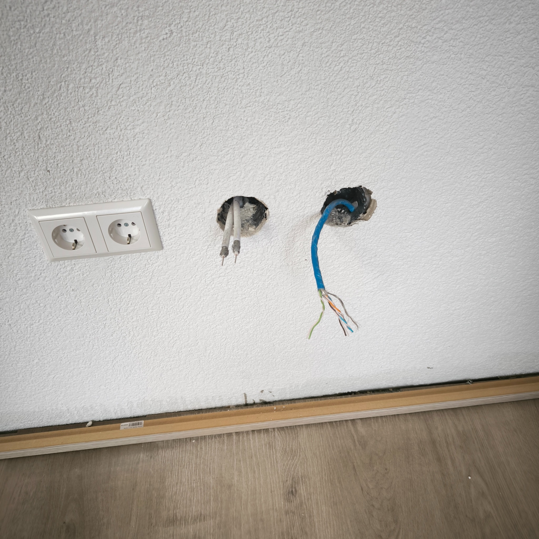 Biała ściana z widoczną podwójną wtyczką oraz dwoma otworami z wystającymi kablami: jeden z przewodami antenowymi, drugi z niebieskim kablem sieciowym z rozplątanymi przewodami w kolorach zielonym,...