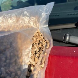 PELTON VALERII SAVCHUK - Pellet drzewny w przezroczystym worku foliowym, leżącym na czerwonej masce samochodu, z widocznym fragmentem srebrnego auta w tle.