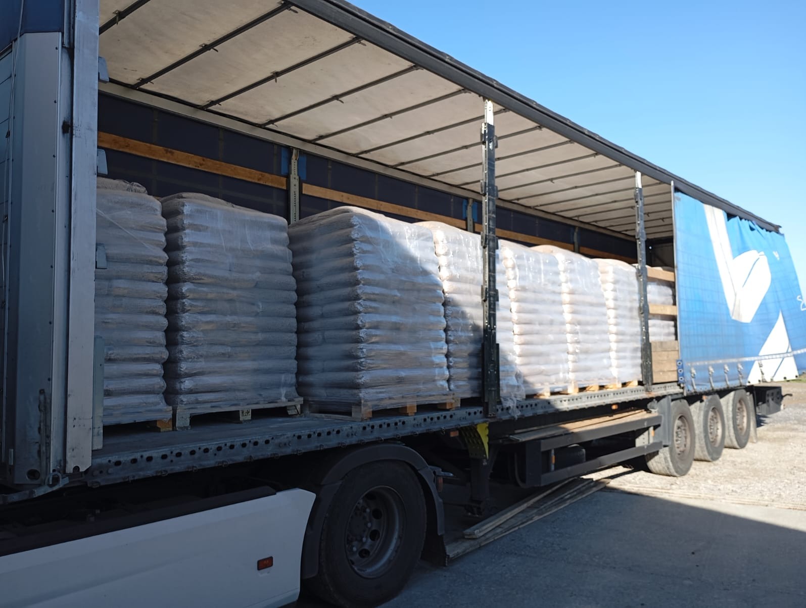 Ciężarówka z naczepą załadowana paletami z workami pelletu, zabezpieczonymi folią stretch, widoczne niebieskie niebo w tle.