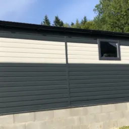 Zewnętrzny widok nowo wybudowanego, dwukolorowego budynku z blachy trapezowej z pojedynczym oknem, osadzonego na betonowej podmurówce, z widocznymi elementami orynnowania i otaczającą zielenią drzew.