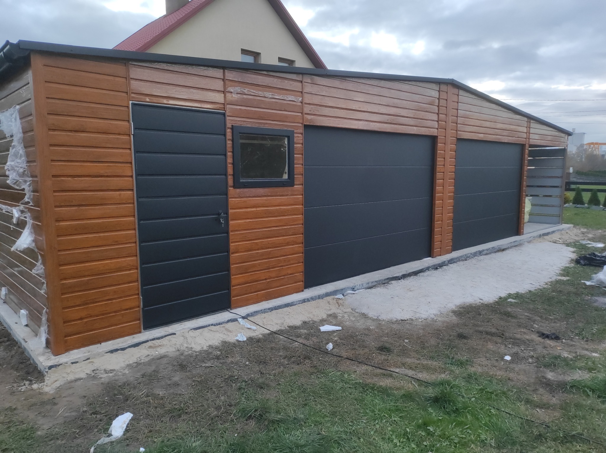 Nowo postawiony garaż z drewnianą elewacją, trzema segmentowymi bramami w kolorze grafitowym i drzwiami bocznymi, usytuowany na trawniku z widocznymi śladami prac budowlanych.