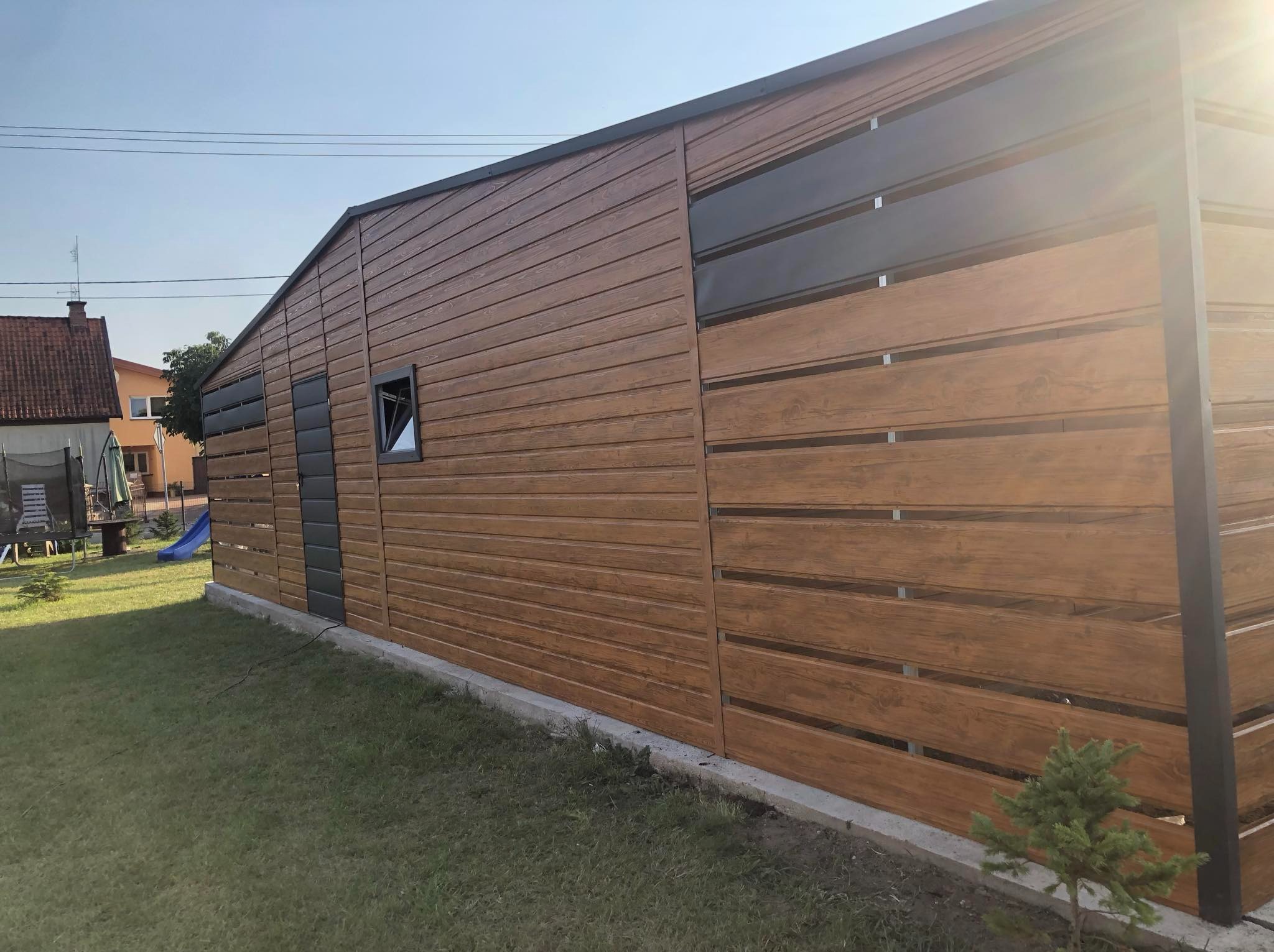 Nowoczesny garaż blaszany z drewnopodobną elewacją i czarnymi akcentami, widok z zewnątrz na tle zielonego trawnika.